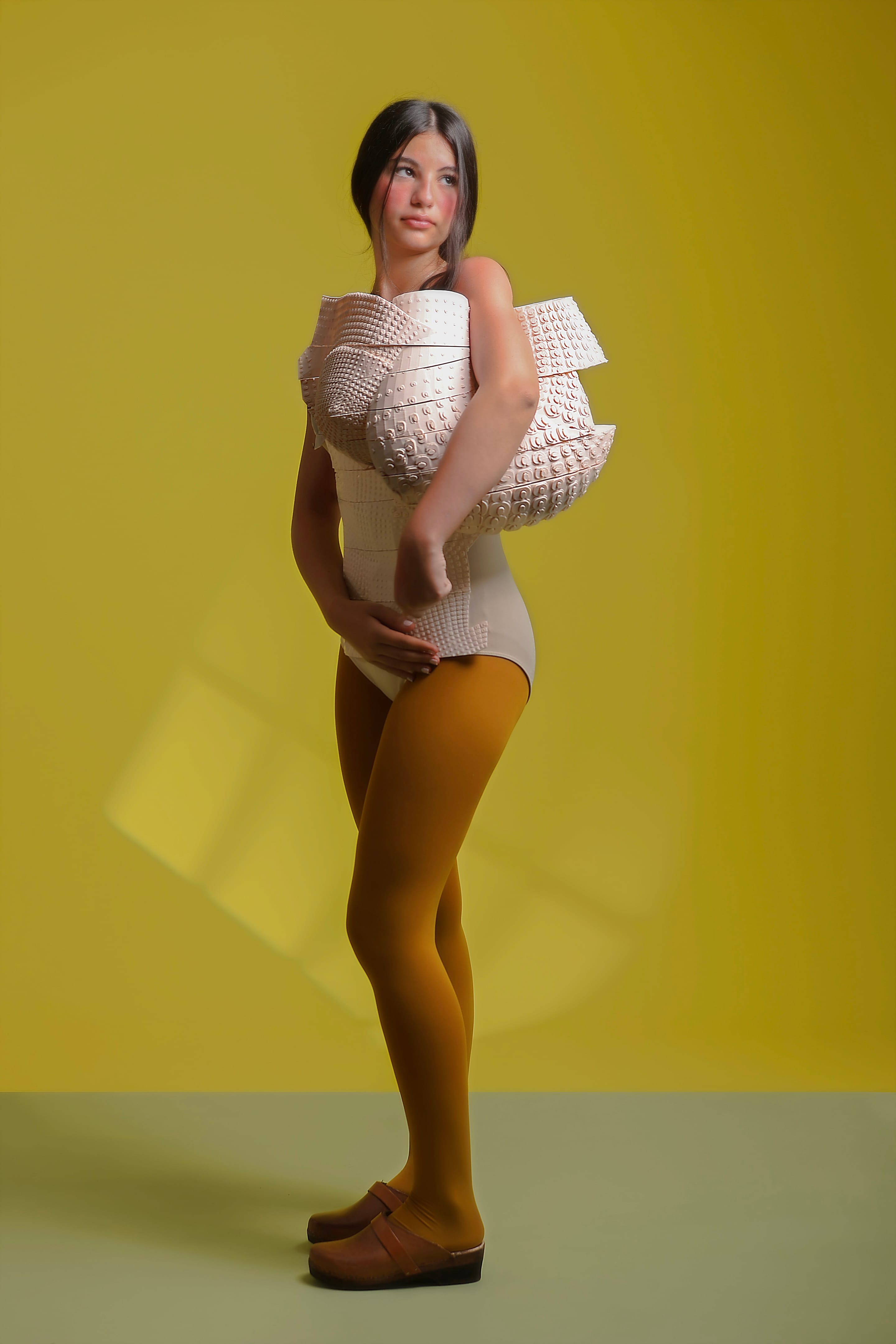 Full-length side view of the model glancing back over the shoulder in the sculpted corset, mustard tights and brown clogs, olive-yellow backdrop