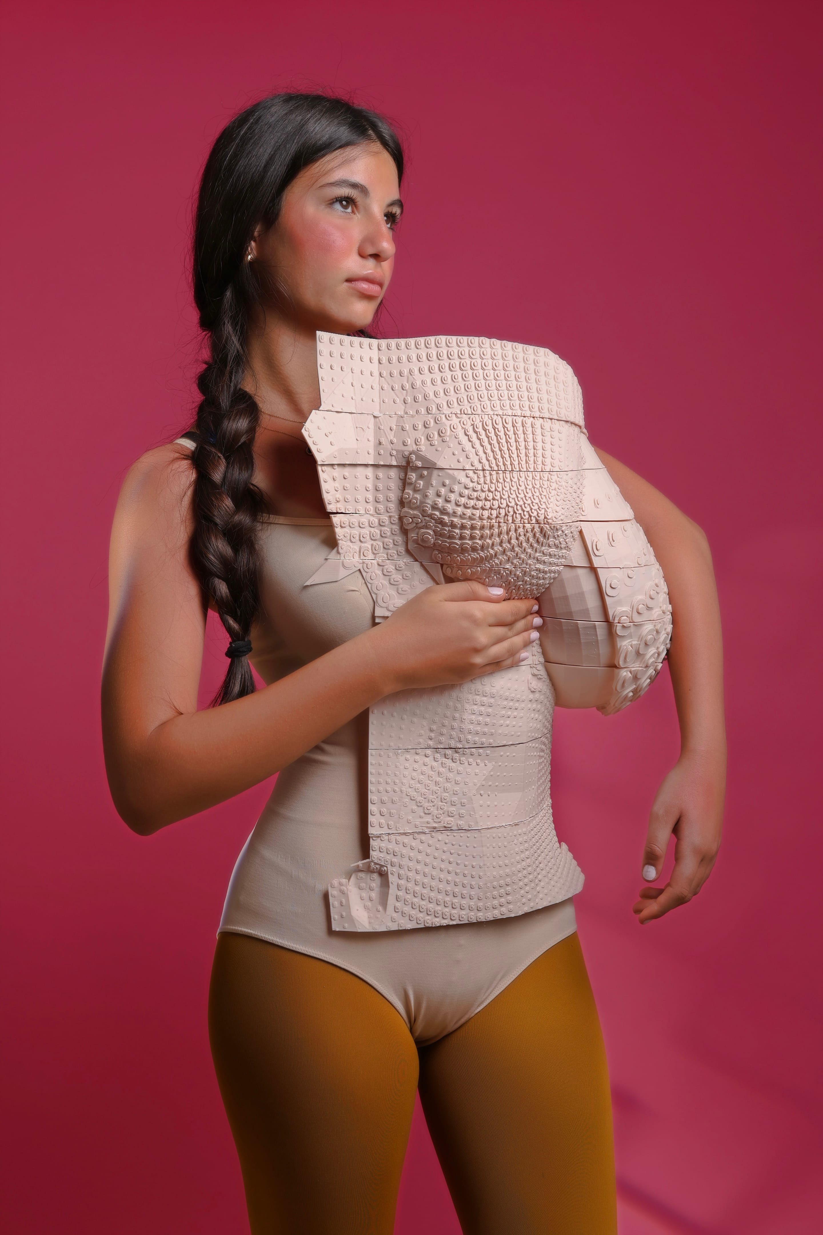Three-quarter-length shot of the model with a braid, one hand resting on the textured corset, mustard tights, magenta backdrop
