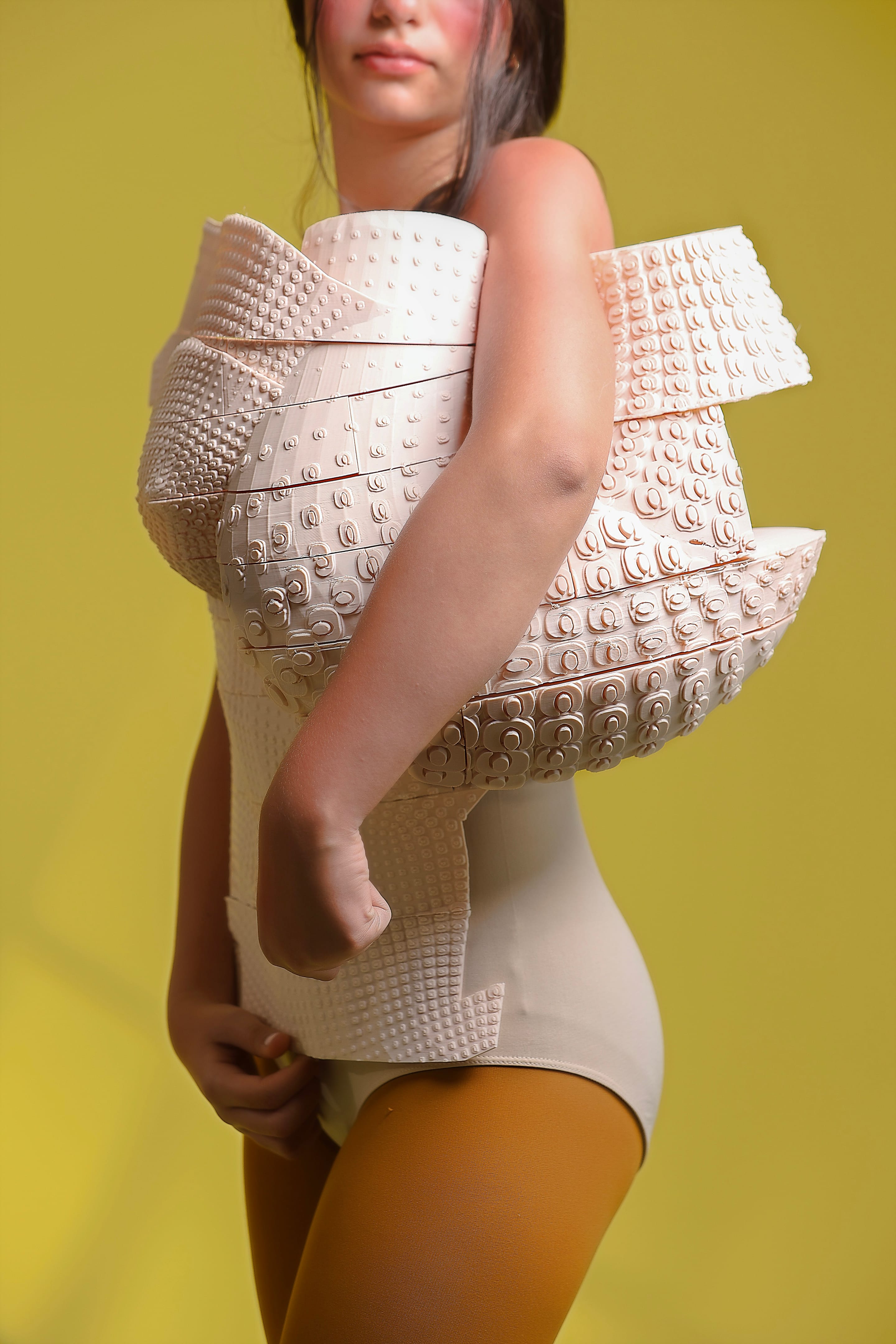 Side close-up of the corset's layered curved bands with raised dot and swirl motifs wrapping the arm, mustard tights, olive-yellow backdrop