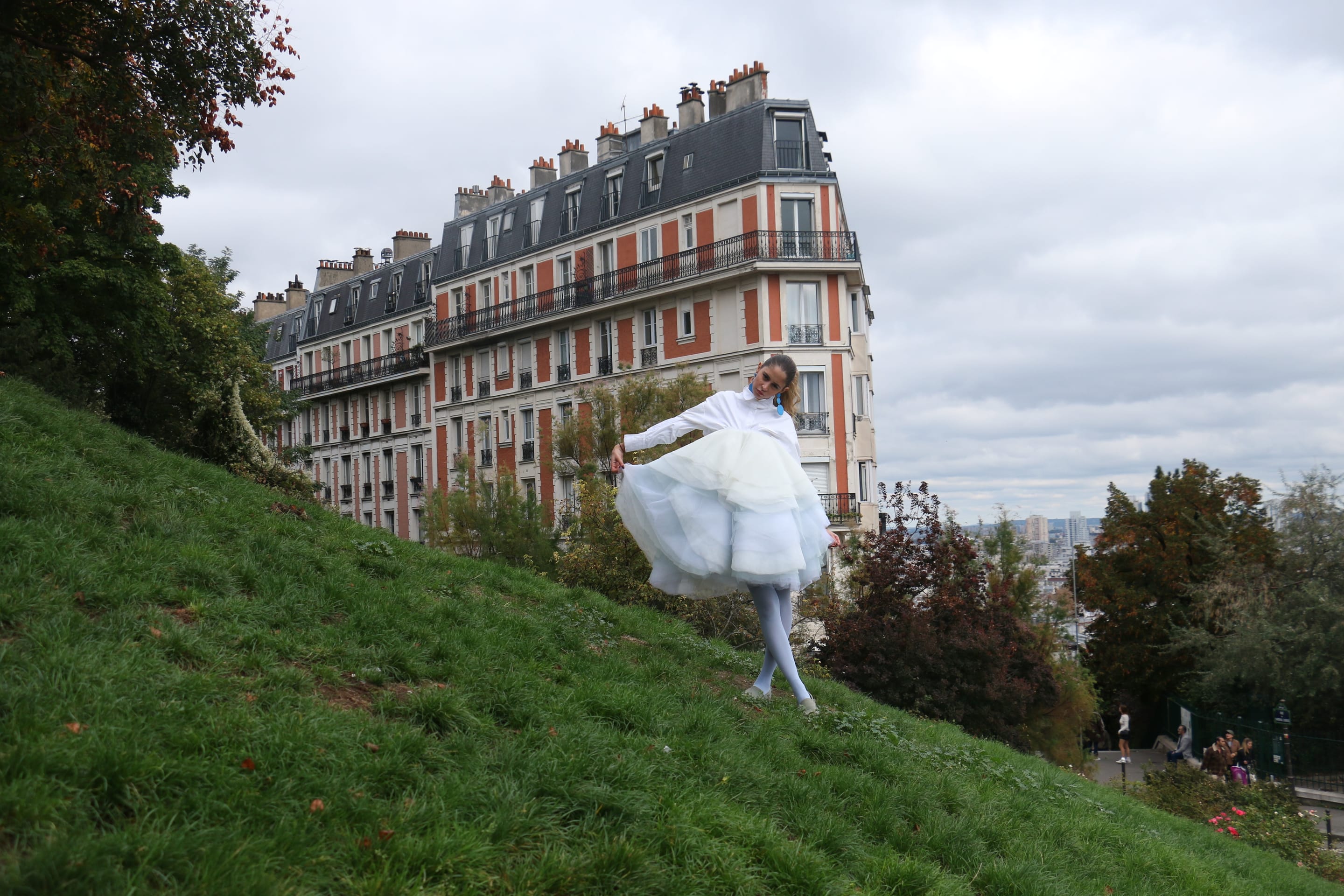 Wide shot of a model in a white shirt and pale blue tulle skirt stepping down a grassy hill, arms out, below Parisian apartments