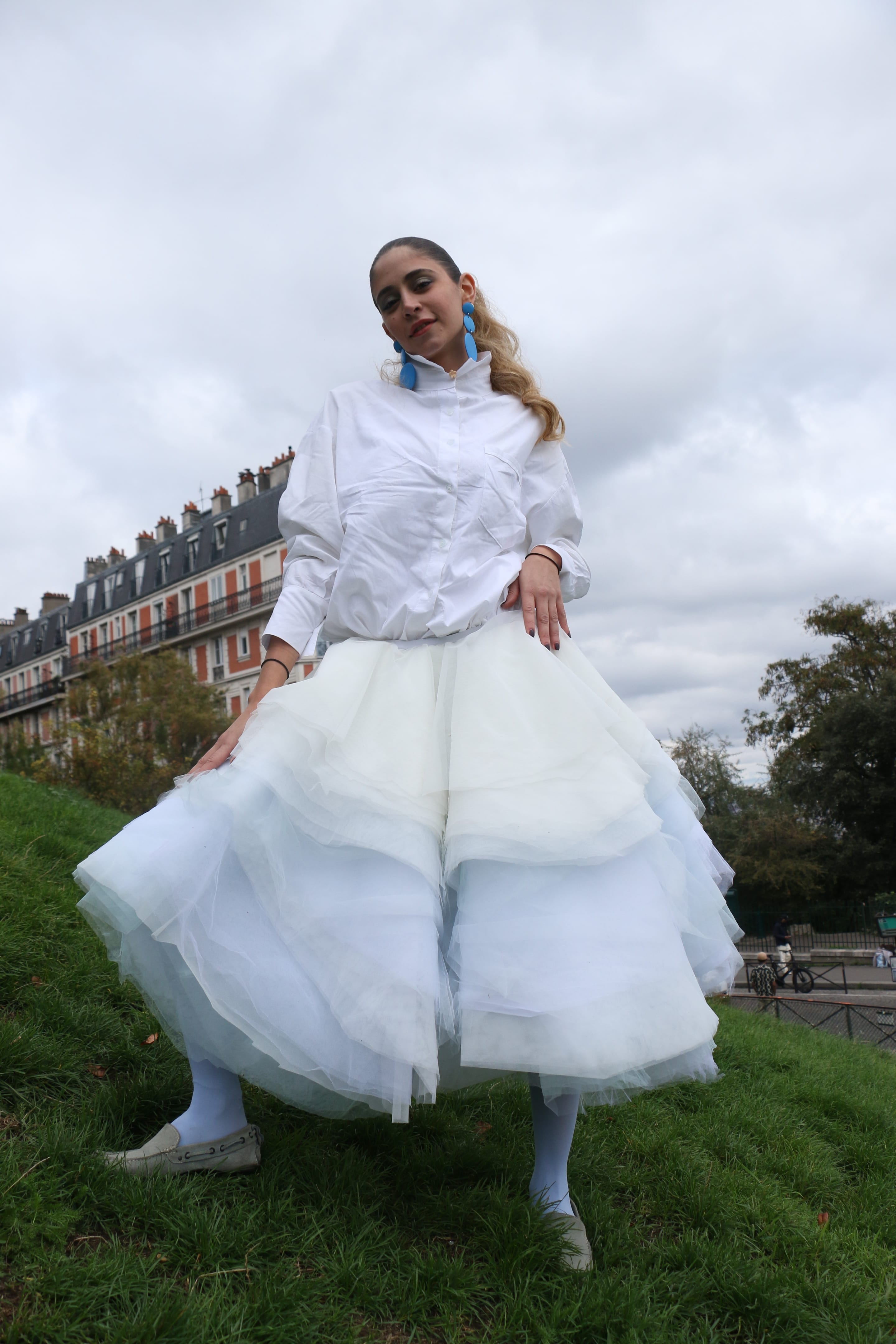Low-angle full-length shot of a model on a grassy slope, hands on hips, in a white shirt and layered pale blue tulle skirt