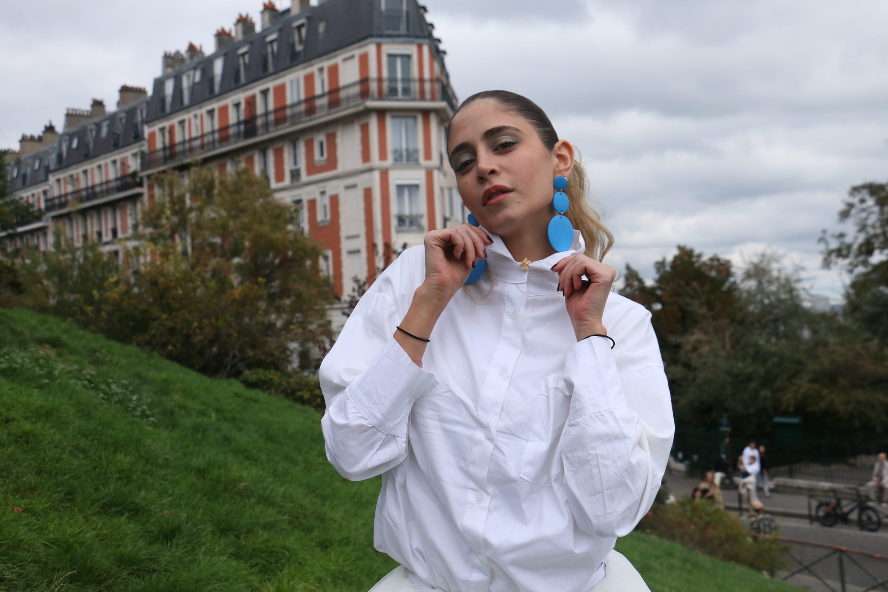 Waist-up portrait of a model holding the collar of an oversized white shirt, wearing large blue disc earrings, apartment building behind