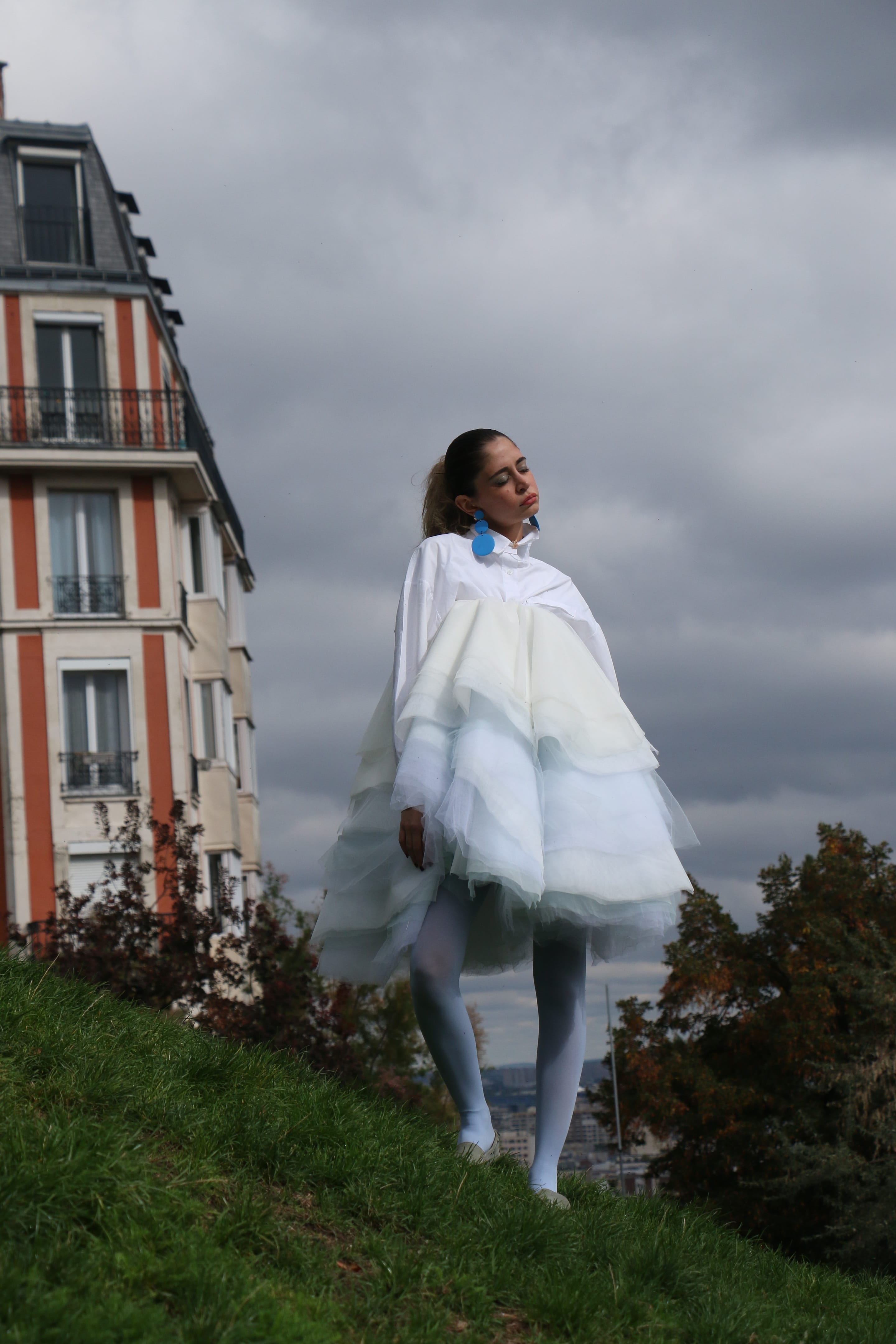 Low-angle shot of a model standing on a grassy slope with eyes closed and chin raised under a dark stormy sky, tulle skirt billowing