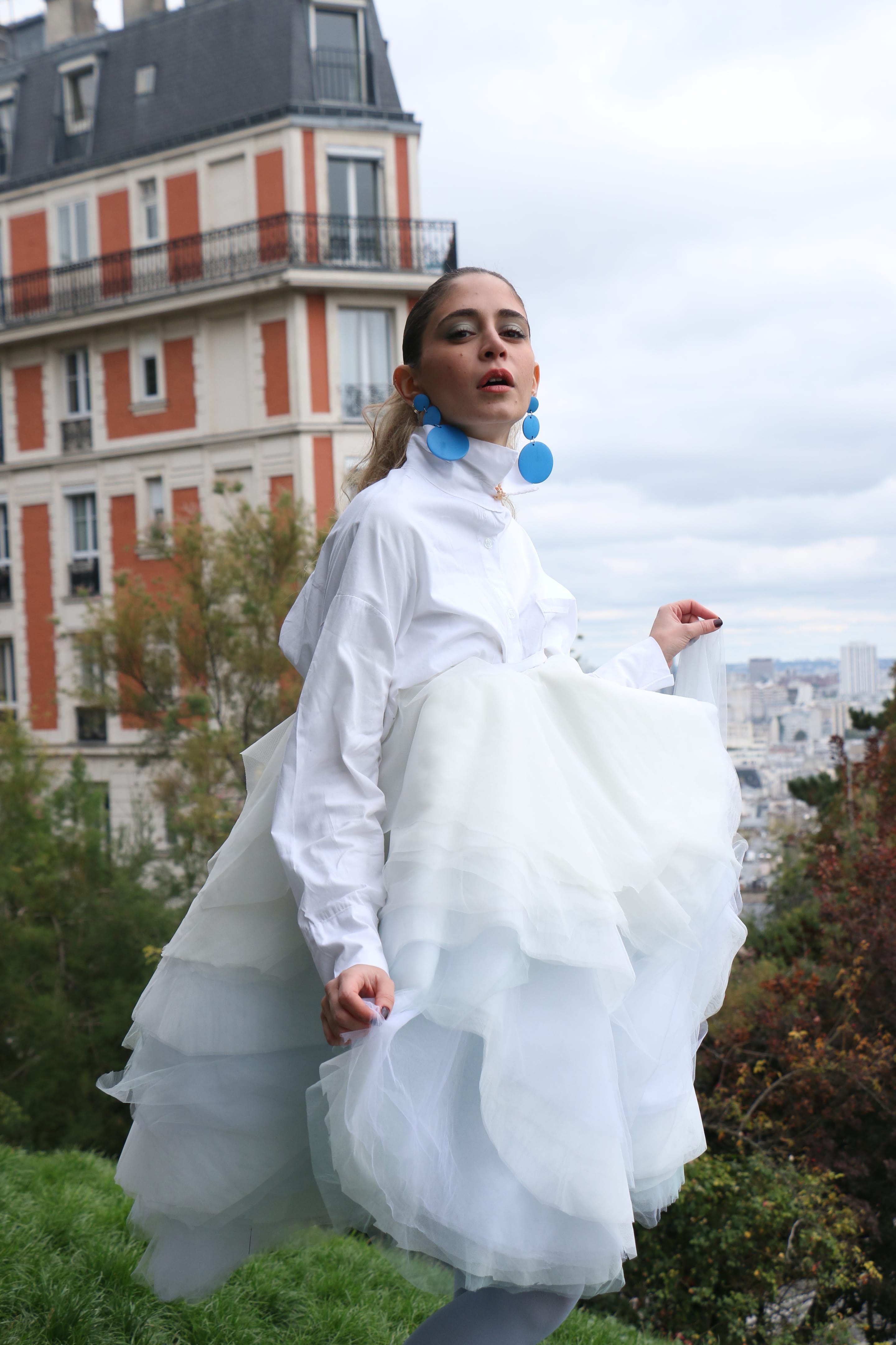 Model in a high-collared white shirt lifting a tiered tulle skirt mid-step and glancing at the camera, city rooftops behind