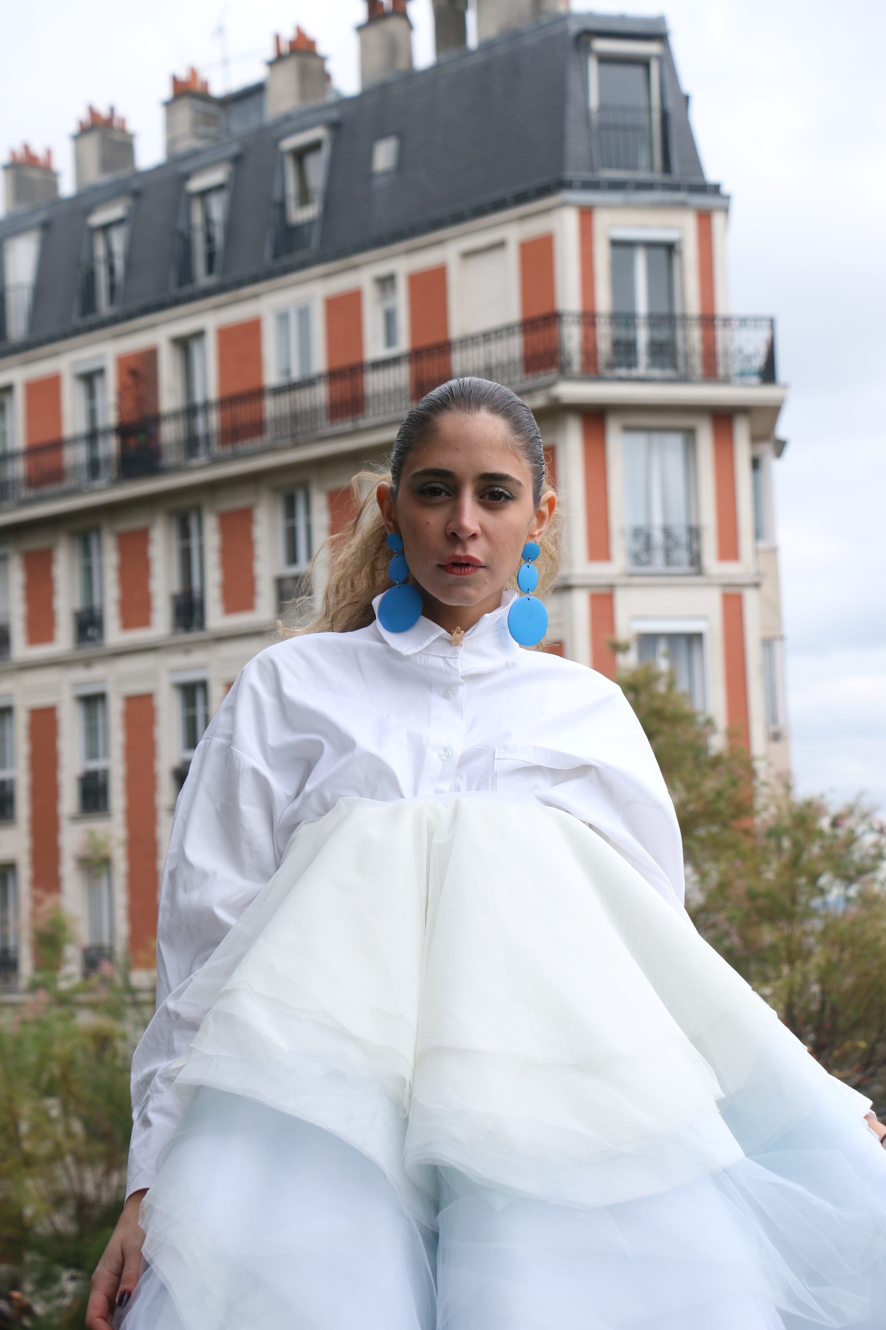 Front-facing portrait of a model with a direct gaze, white shirt gathered over a voluminous tulle skirt, blue disc earrings, apartment building behind