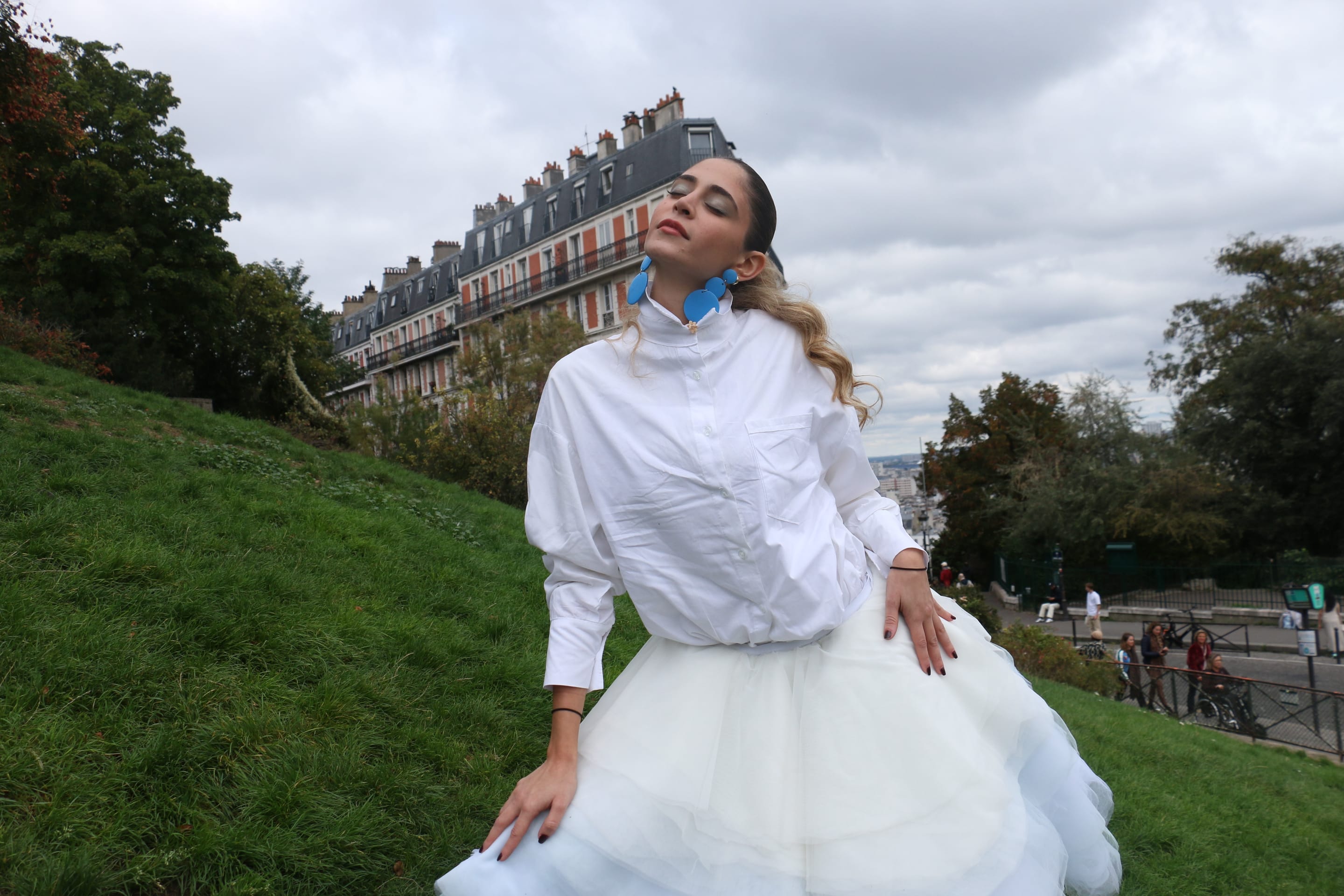 Model sitting low on a grassy hillside, head tilted back and eyes closed, one hand on the hip, in a white shirt and tulle skirt
