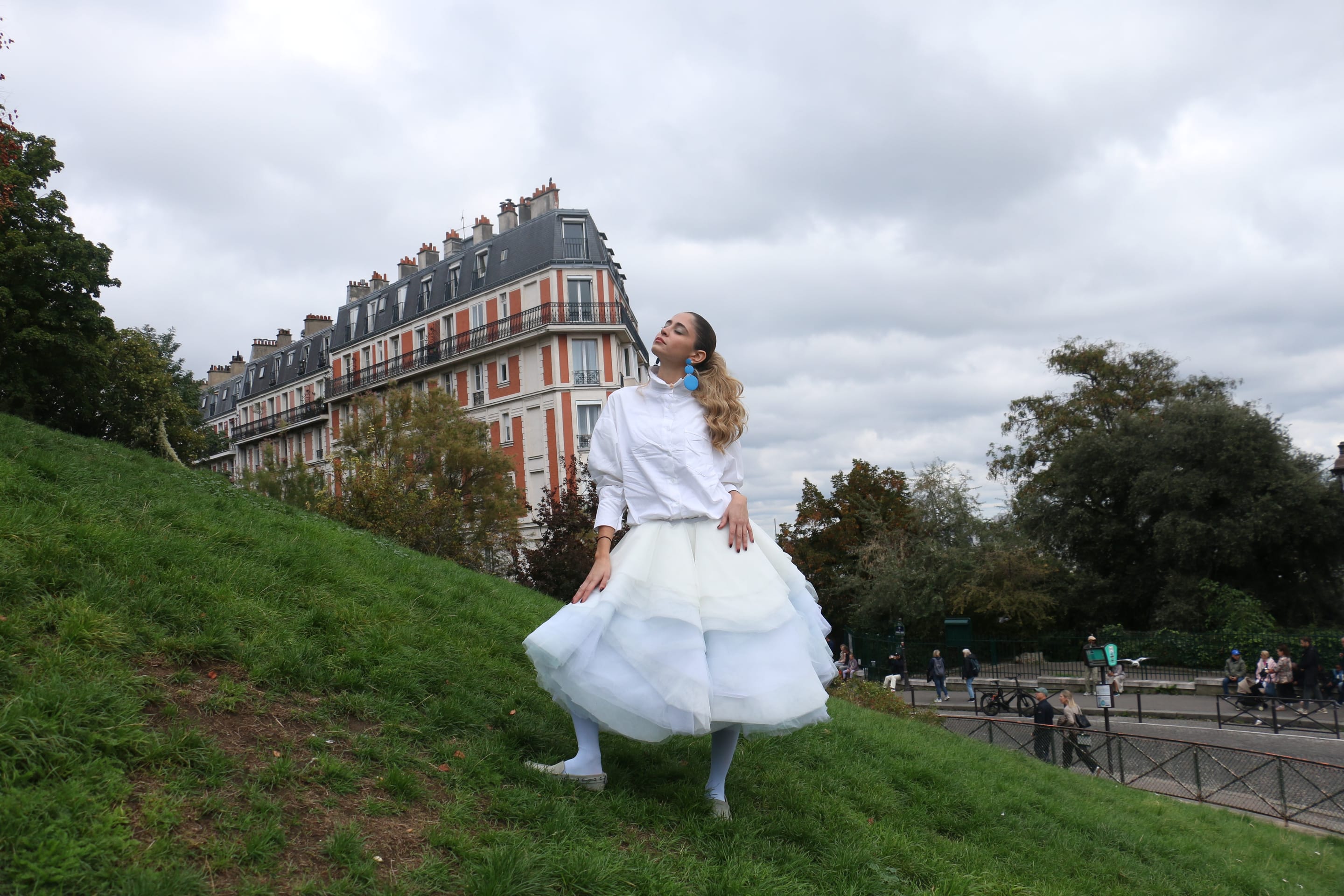 Full-length shot of a model standing on a grassy slope with head turned aside, hands resting on the tulle skirt, park railing below