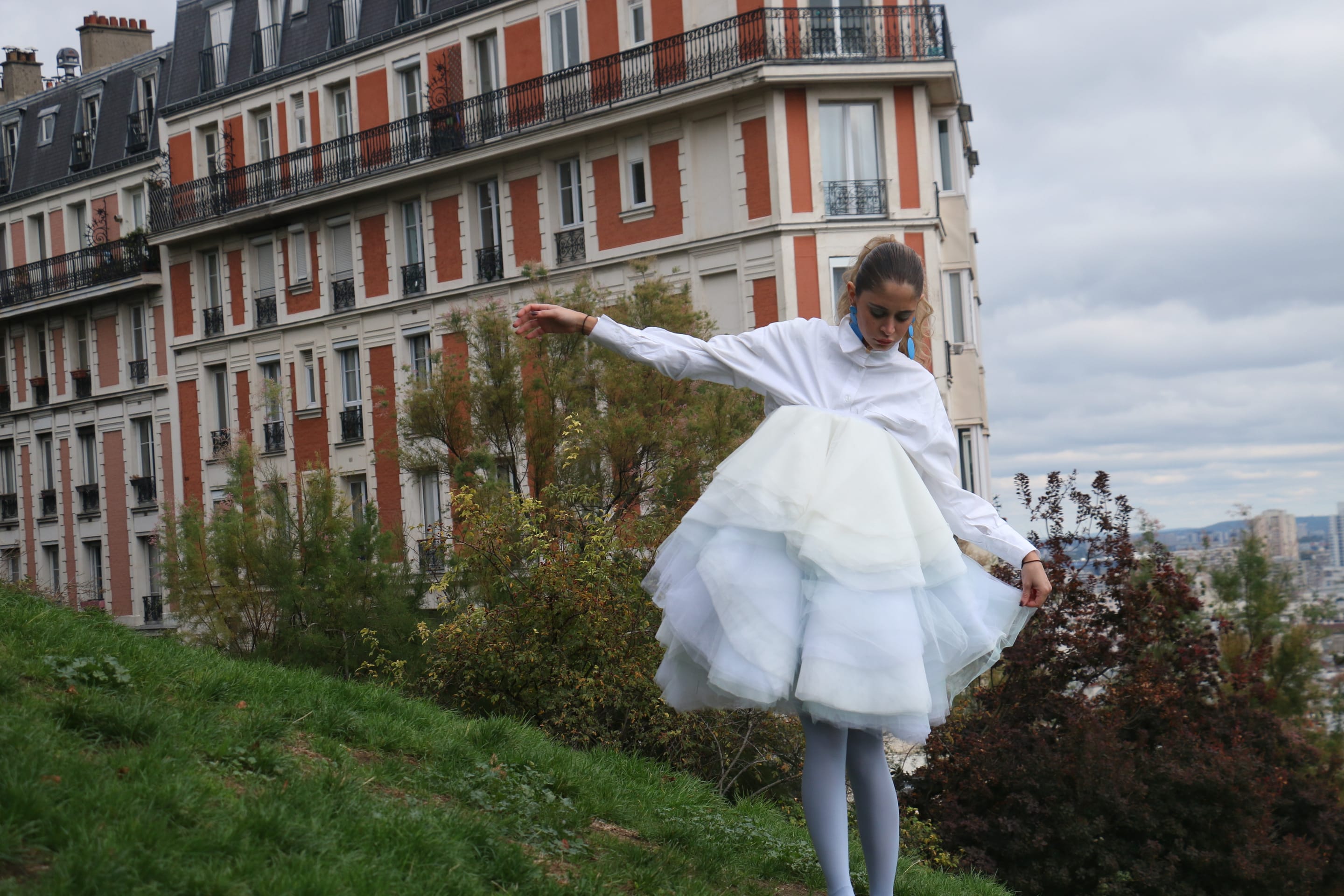 Model in a white shirt balancing with arms spread wide, looking down at the grassy slope as the tulle skirt flares, apartment facade behind