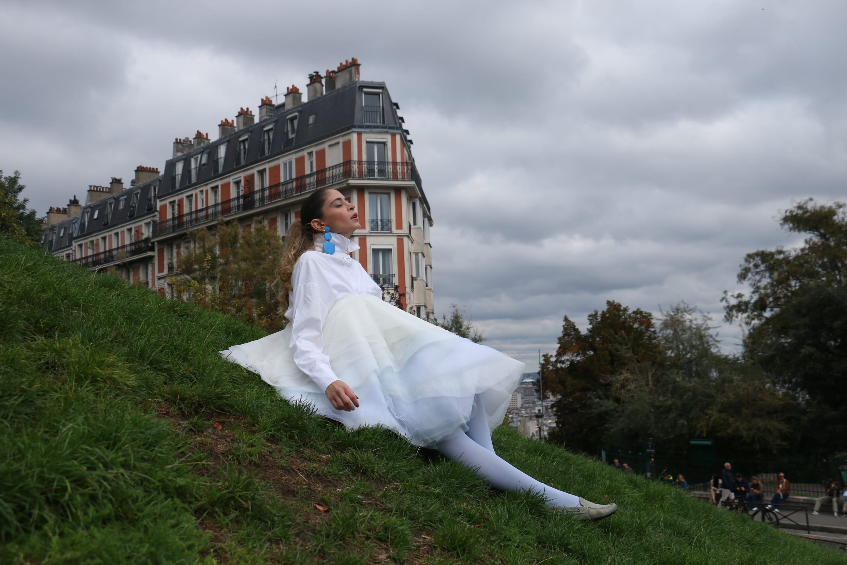 Model reclining on a steep grassy slope with eyes closed and face turned up to a cloudy sky, tulle skirt spread across the grass