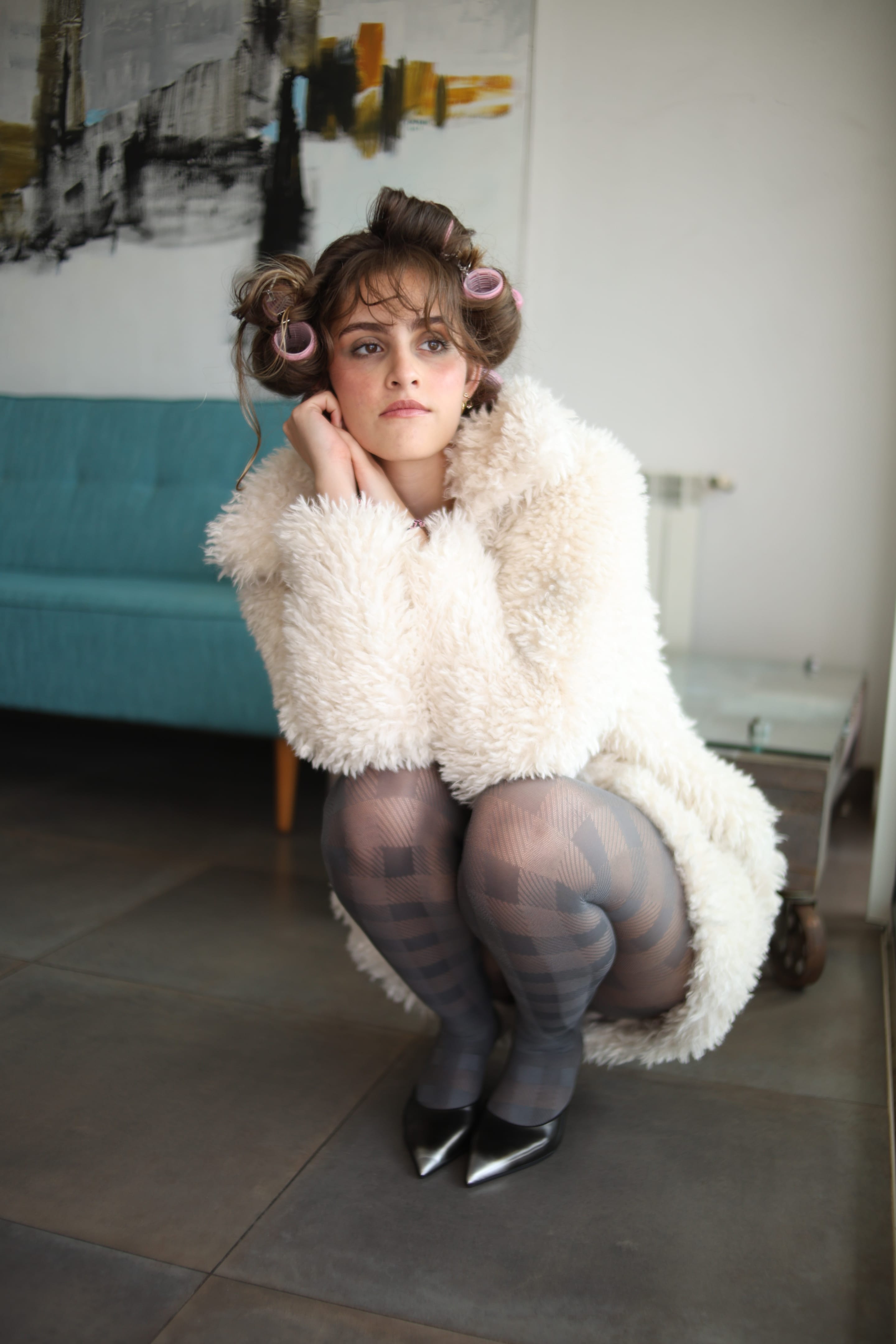 Model in hair rollers and a shaggy cream coat crouching on a tiled floor, chin resting on one hand, teal sofa behind