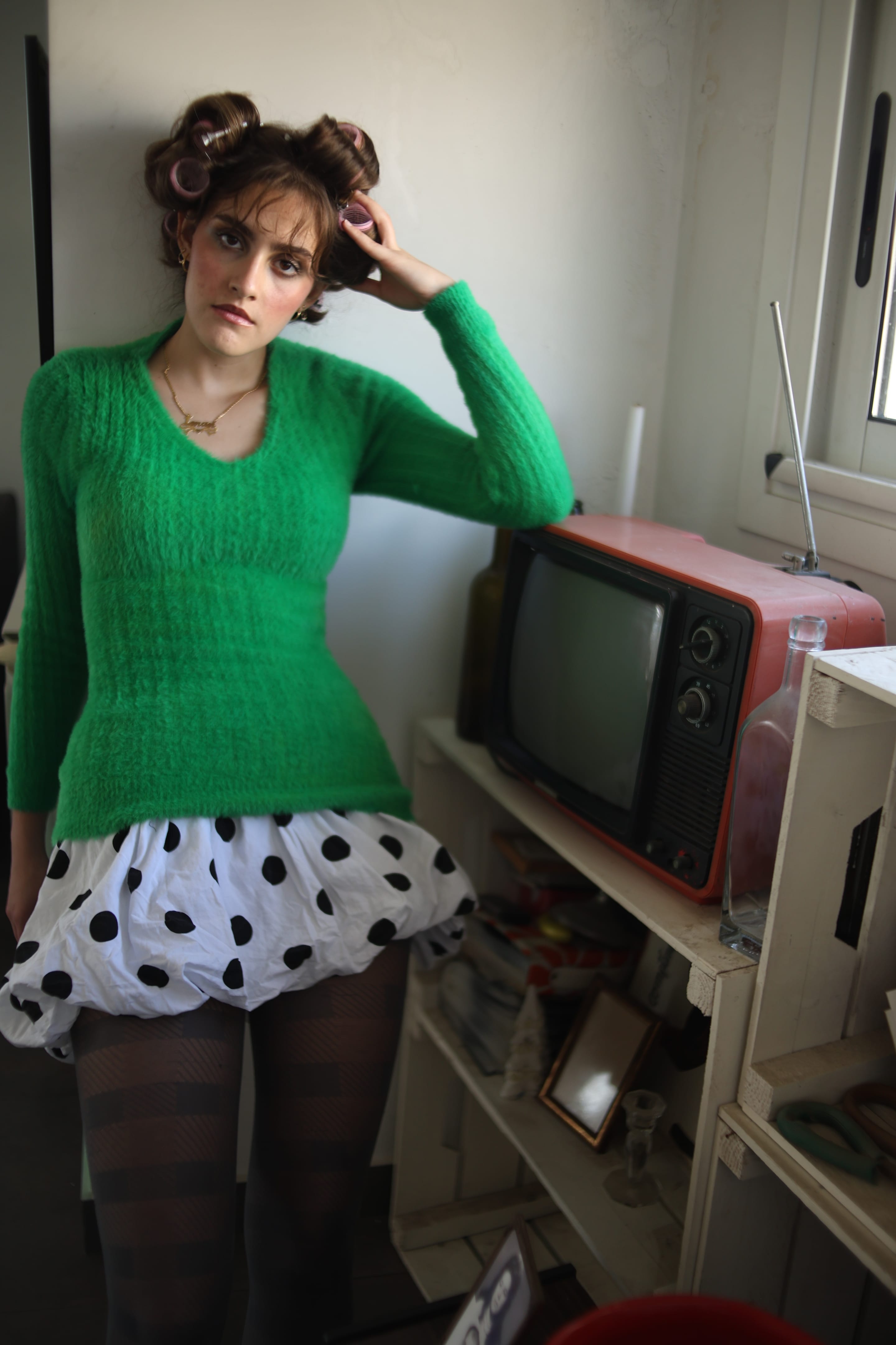 Model in a green sweater and polka-dot bubble skirt, one hand at the hair rollers, standing beside a vintage orange-red television on crate shelves