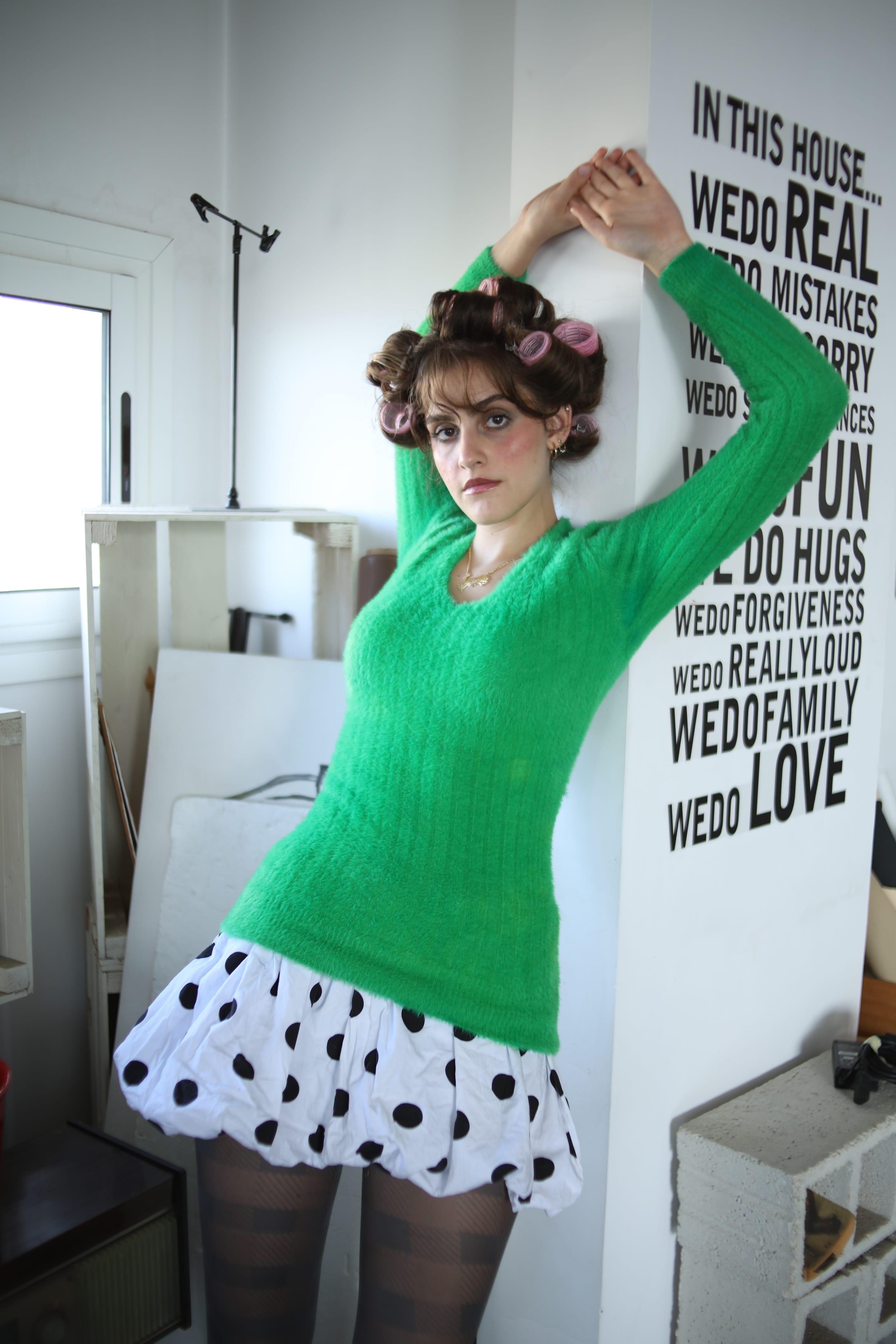 Model in hair rollers, a fuzzy green sweater and black-and-white polka-dot bubble skirt, arms raised against a wall lettered with slogans