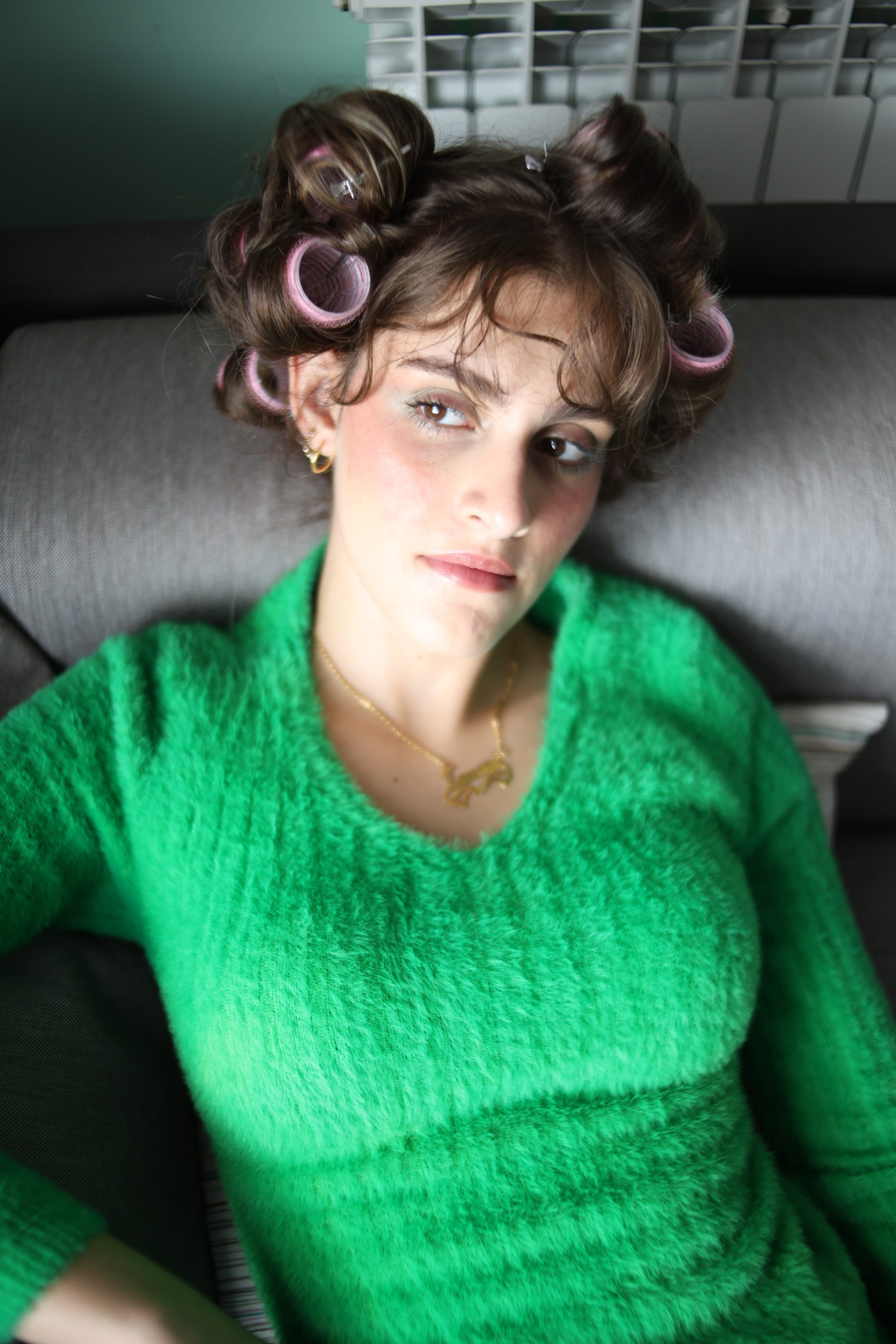 Close-up from above of a model in pink hair rollers and a fuzzy green sweater reclining on a grey sofa, a gold necklace at the throat