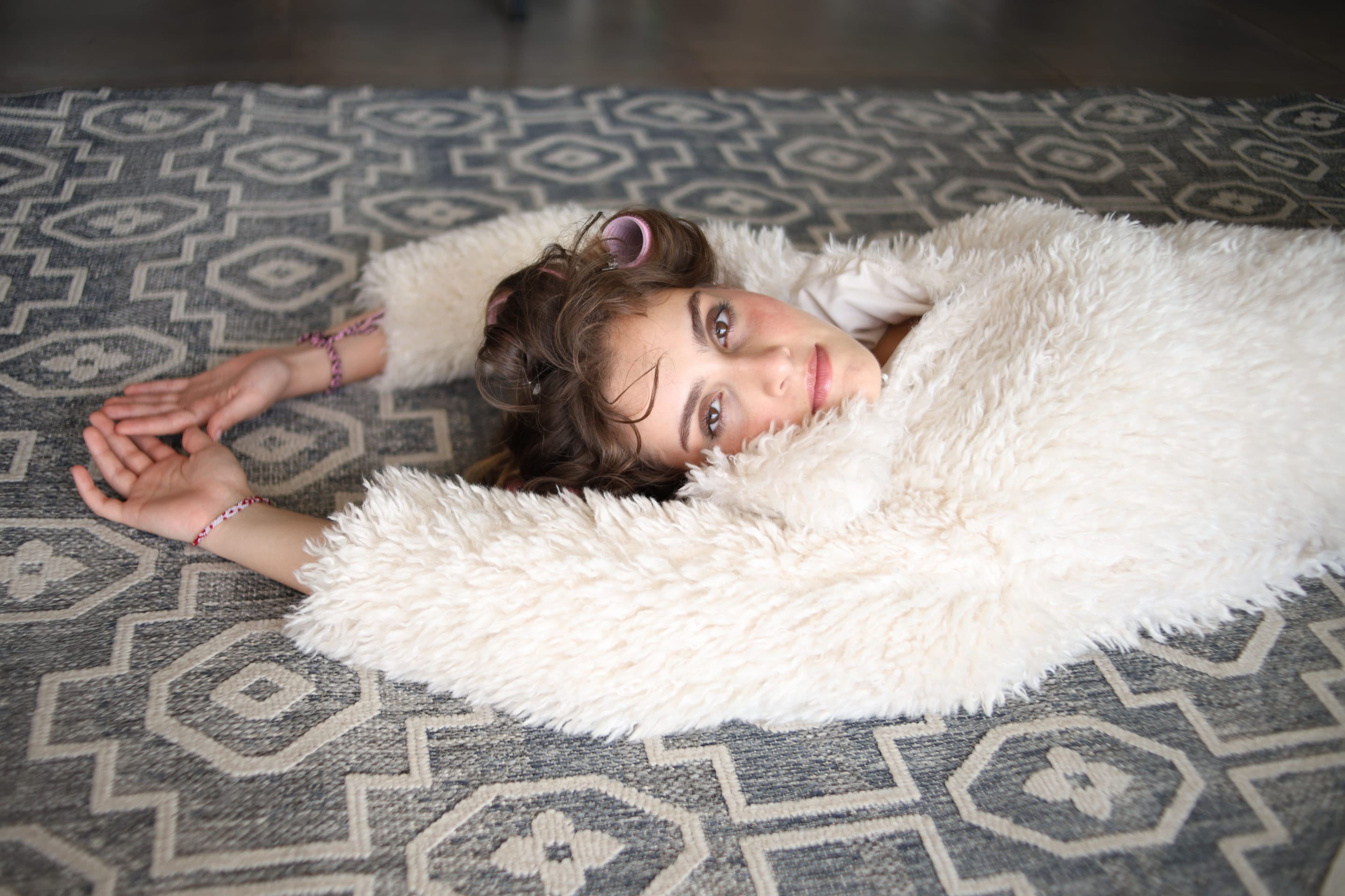 Model lying face up on a grey patterned rug, arms stretched overhead, wrapped in a shaggy cream coat and gazing at the camera