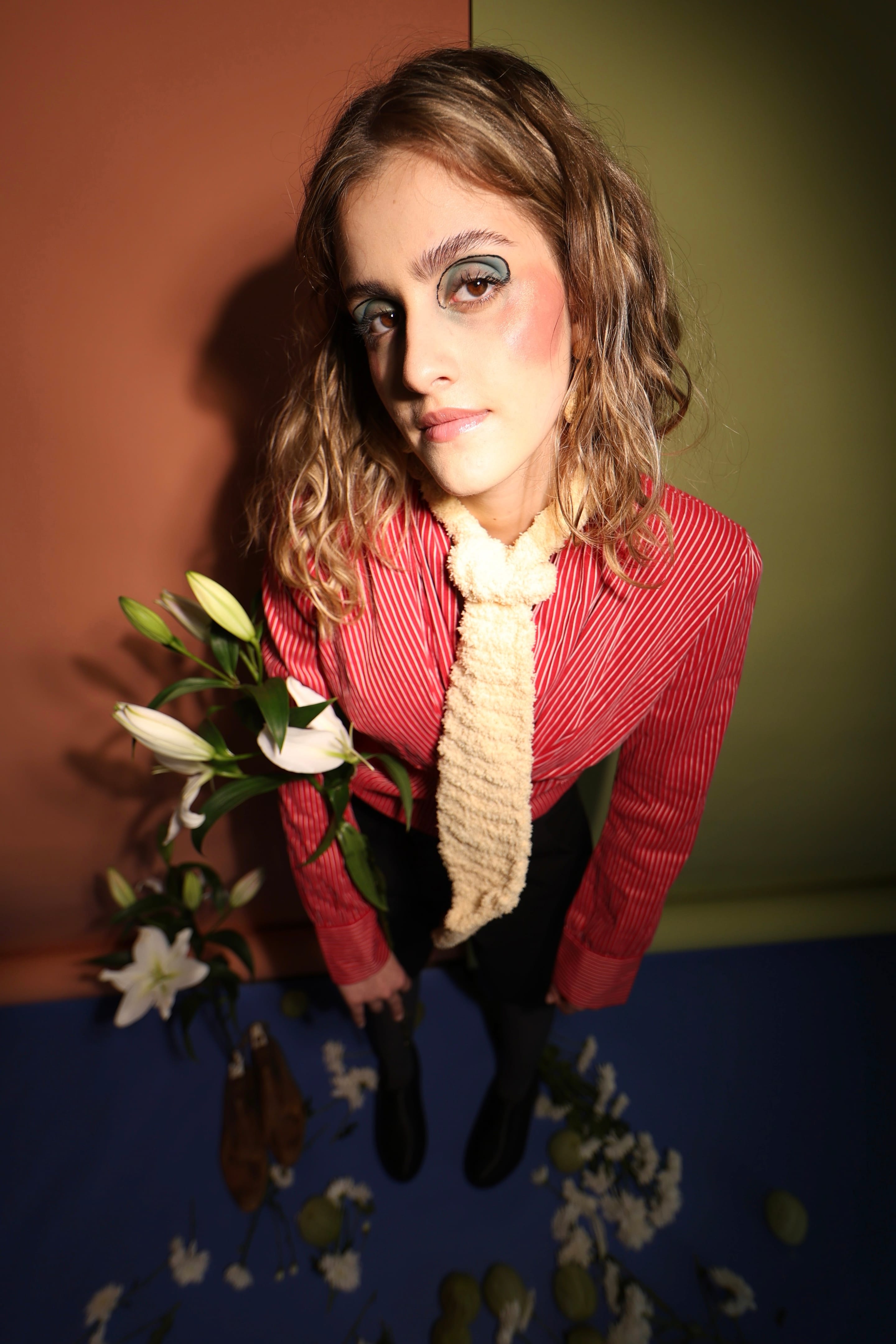 Wide-angle shot from above of a model in a red pinstriped shirt and cream knitted tie, white lilies in the foreground, terracotta and olive backdrop