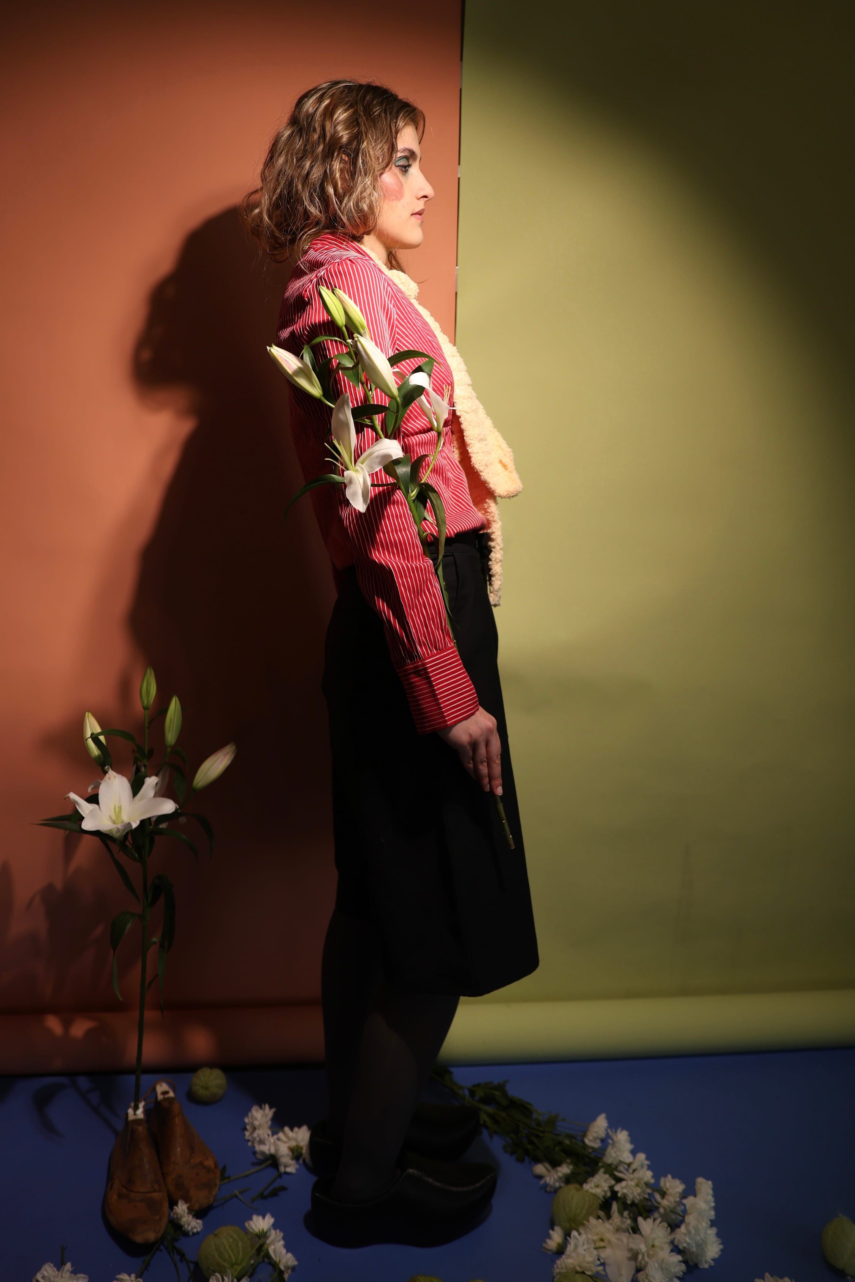 Full-length profile of a model in a red striped shirt and black skirt, a lily tucked at the elbow, against terracotta and olive backdrops