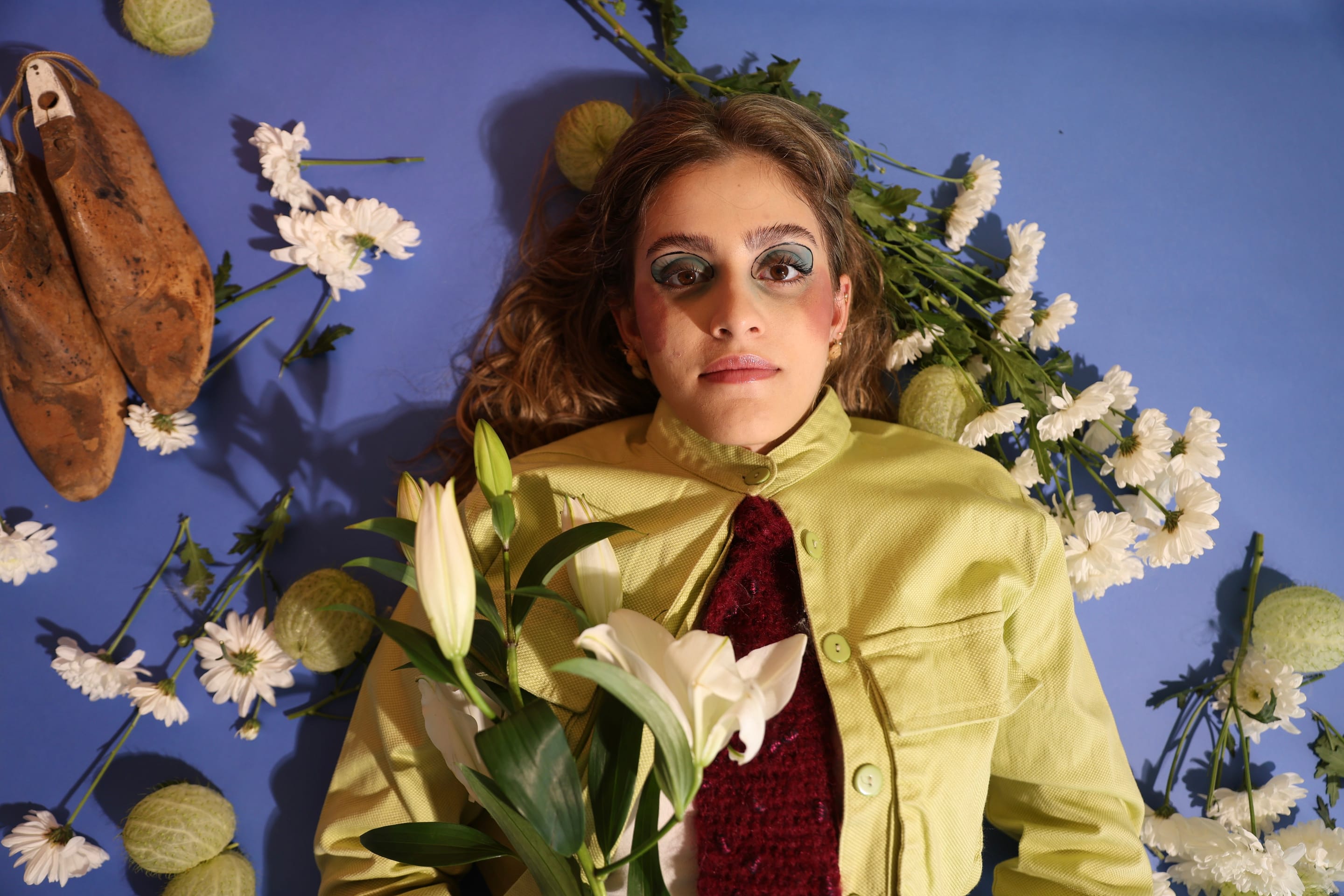 Overhead shot of a model lying in a lime-green jacket and burgundy tie, lilies on the chest, wooden shoe lasts and chrysanthemums all around