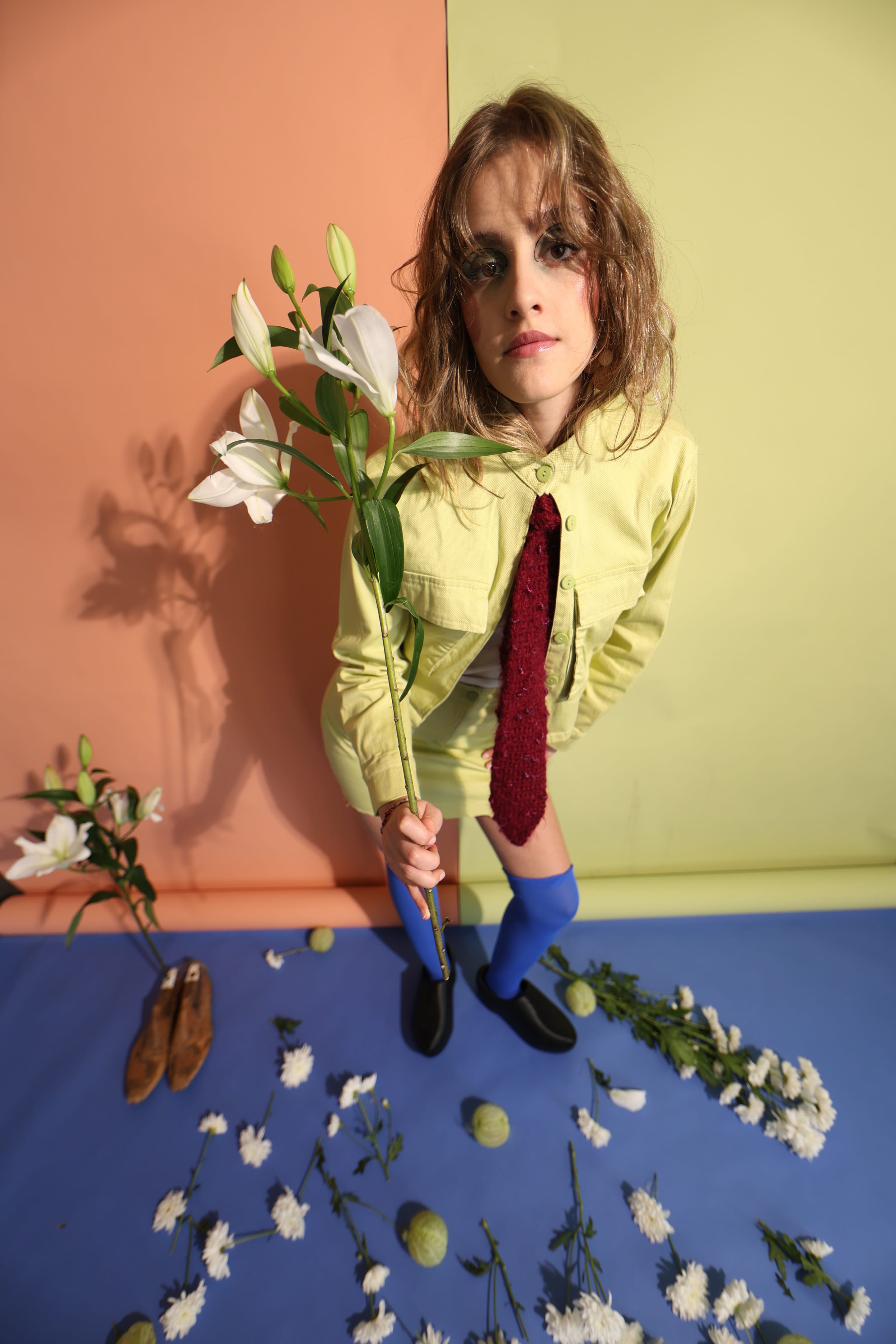 Wide-angle full-length shot of a model leaning towards the camera in a lime-green jacket and skirt, burgundy tie and blue knee socks, holding lilies