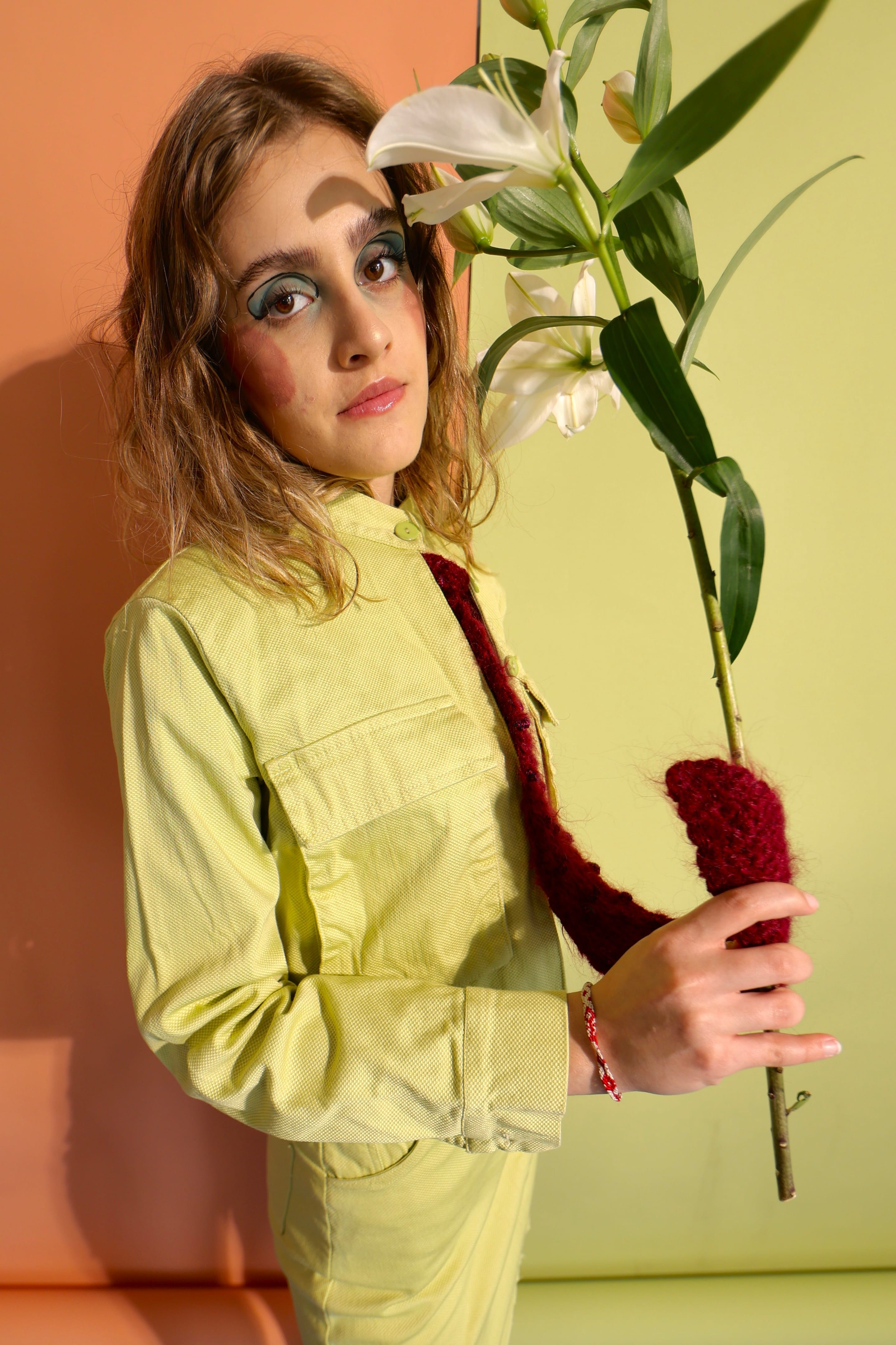 Model turned three-quarters in a lime-green jacket, holding a lily stem and the end of a burgundy knitted tie, blossoms beside the face