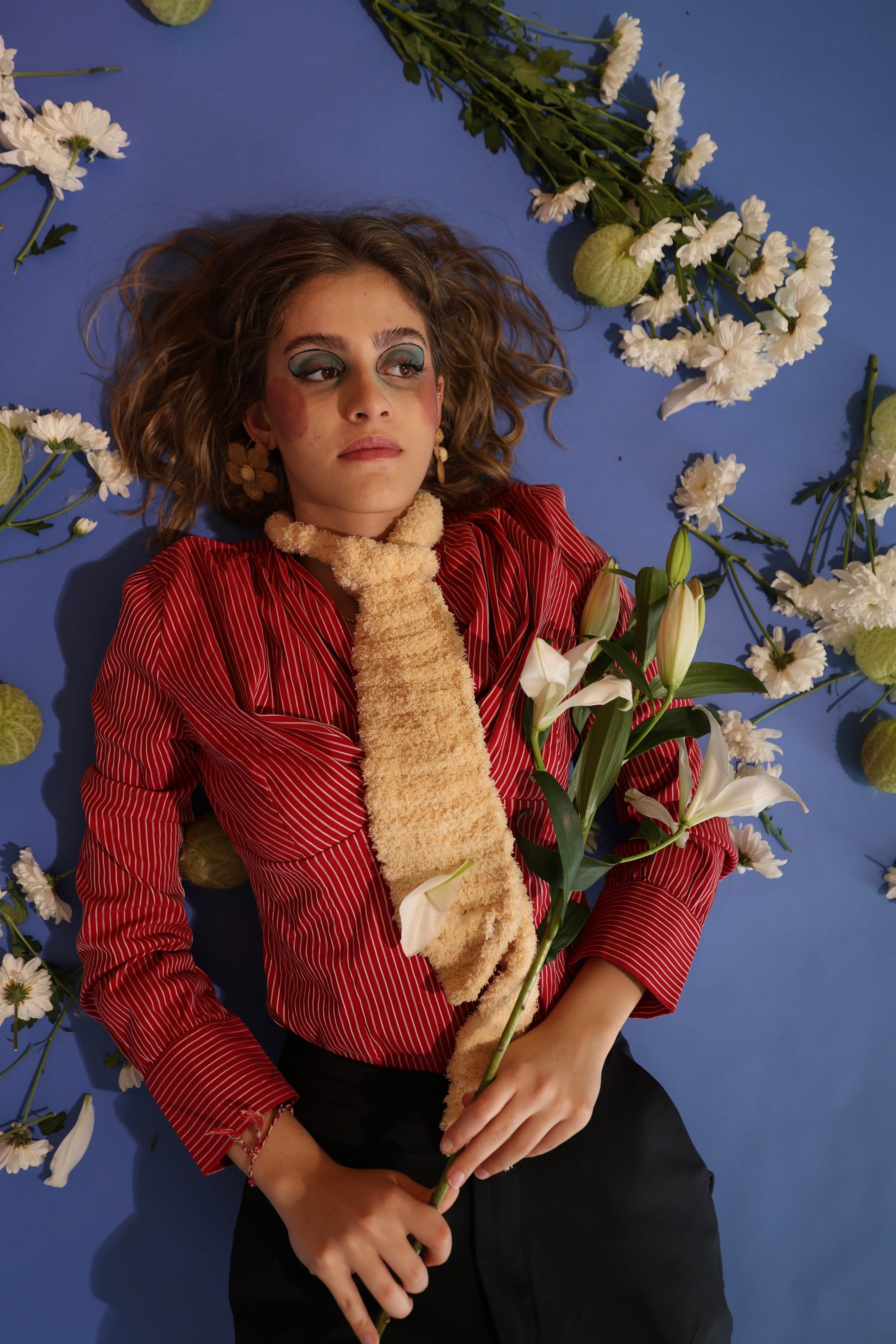 Overhead shot of a model lying on a blue floor in a red striped shirt and cream knitted tie, holding lilies among scattered white chrysanthemums