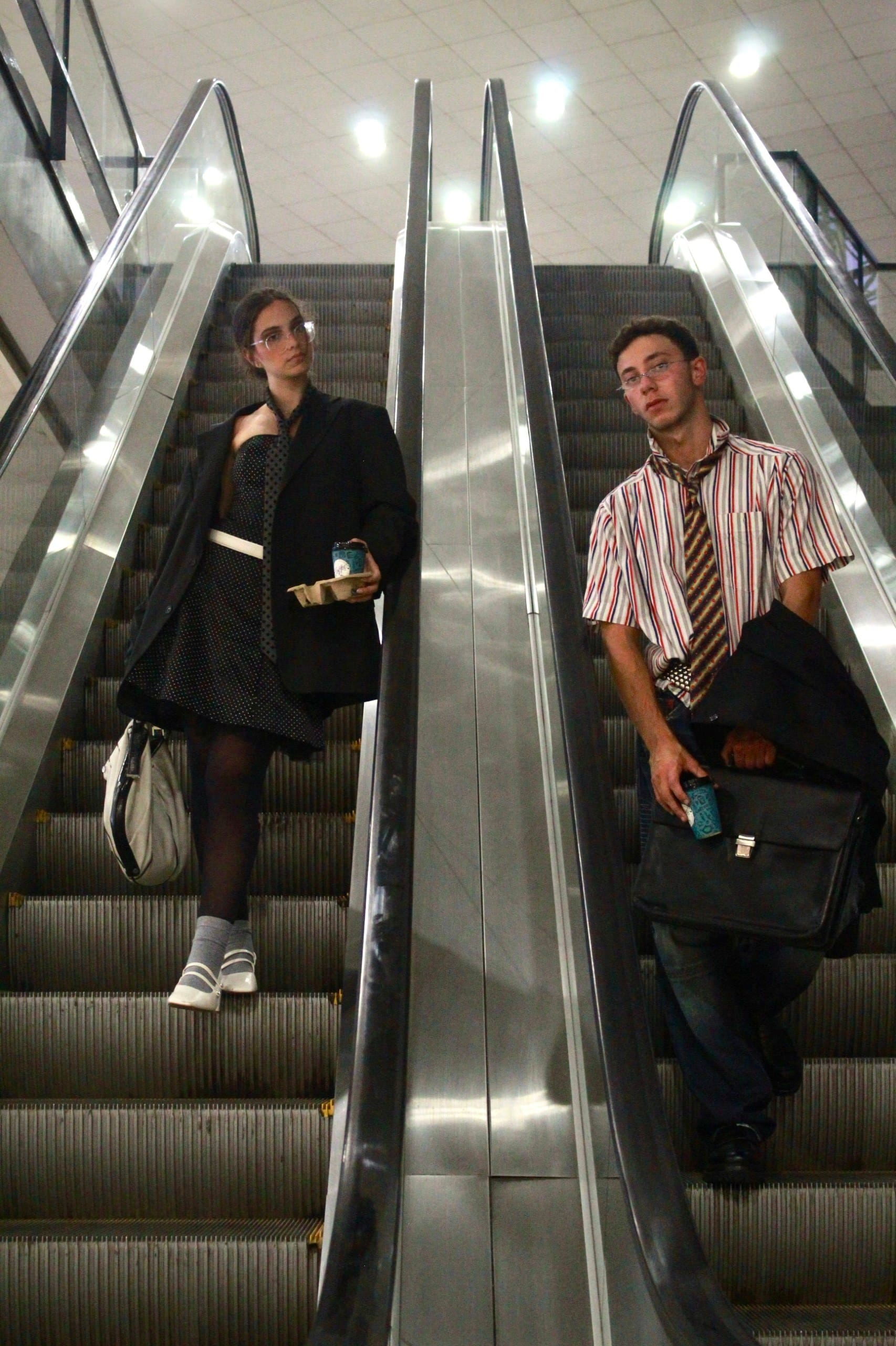 Two models descending side-by-side escalators, one in a blazer and polka-dot dress holding coffee, one in a striped shirt and tie with a briefcase
