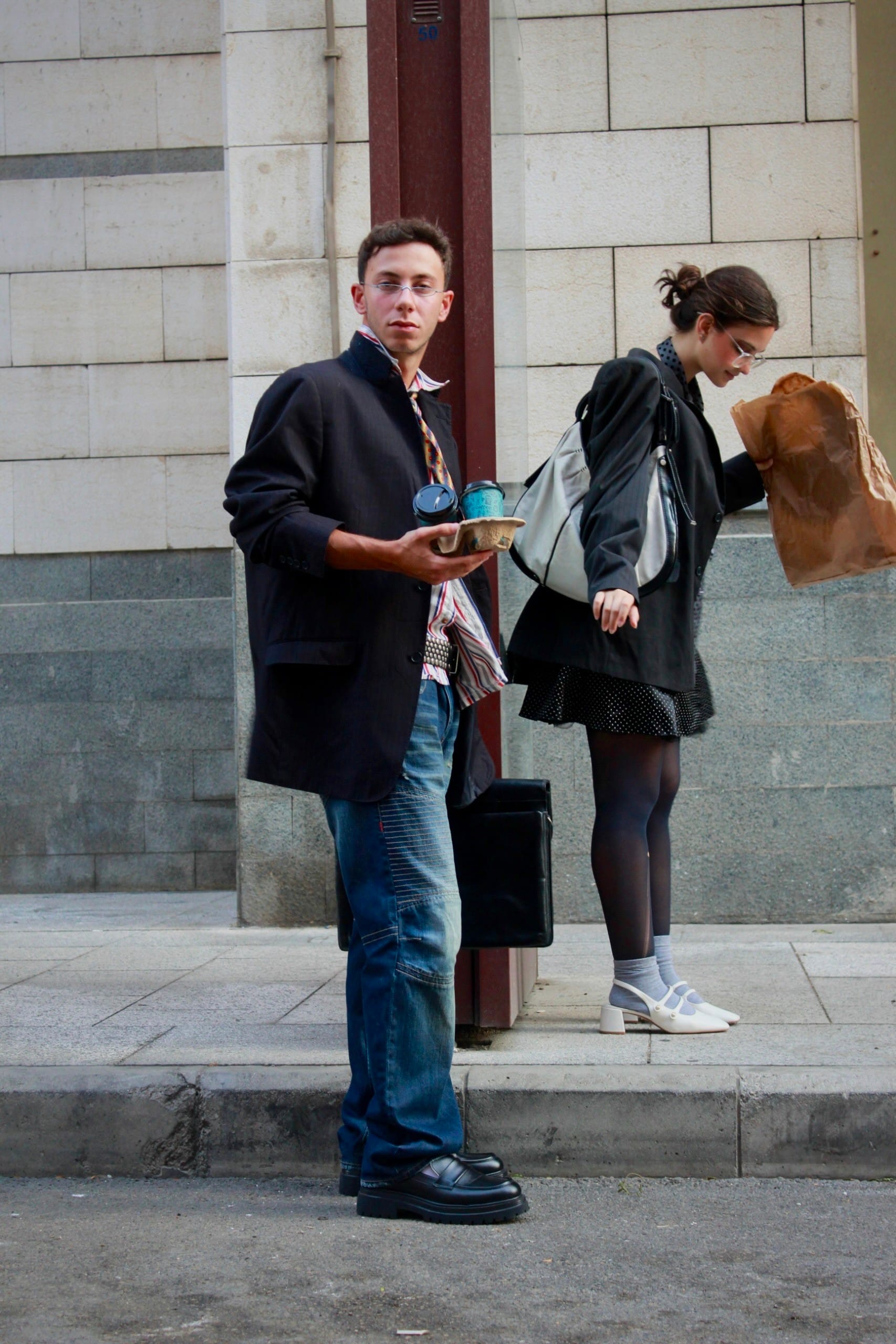 Model in a dark blazer and jeans holding coffees at the kerb while a second model behind peers into a paper bag