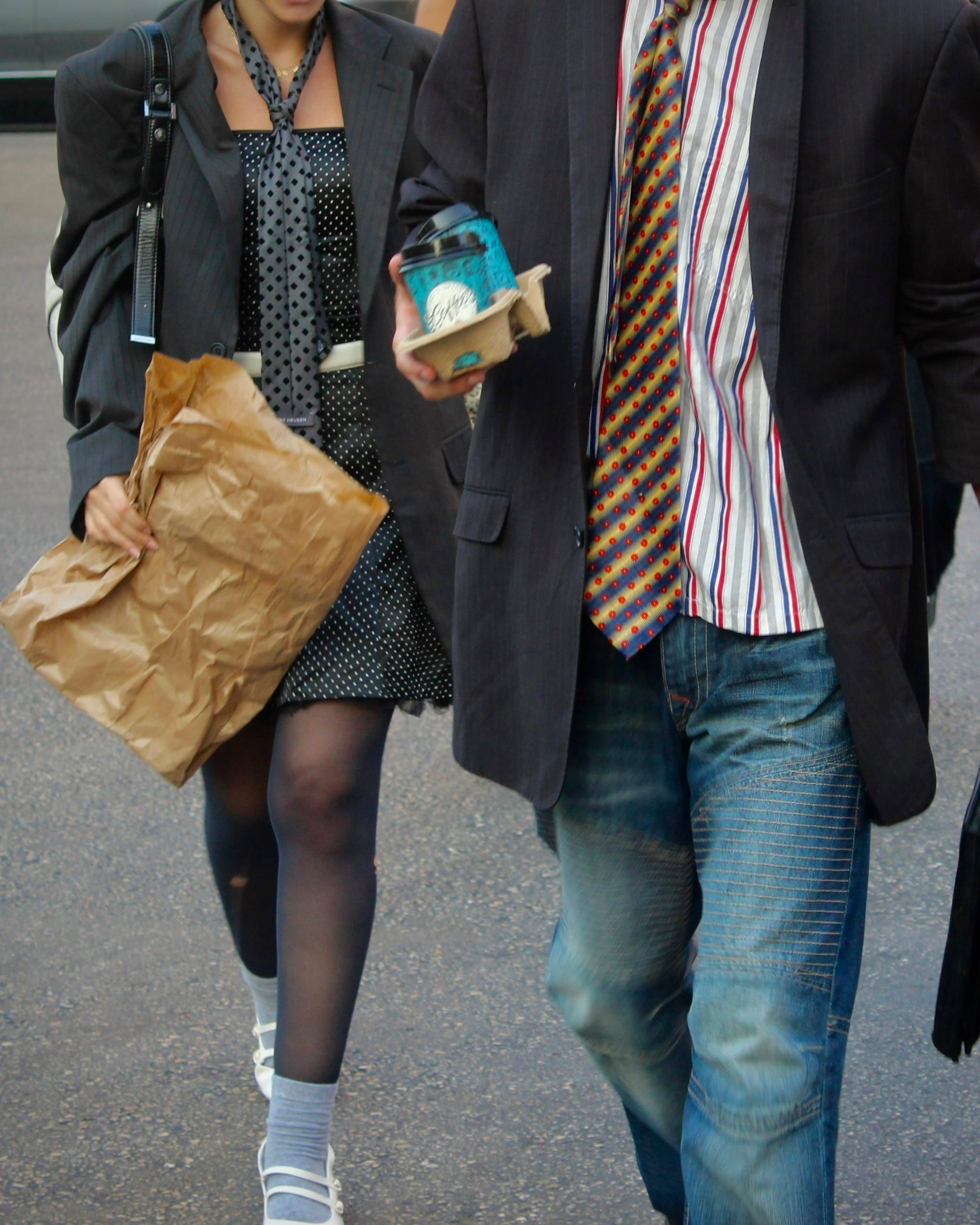 Cropped torsos of two models walking side by side, one in a polka-dot dress carrying a paper bag, one in a pinstripe blazer holding coffees