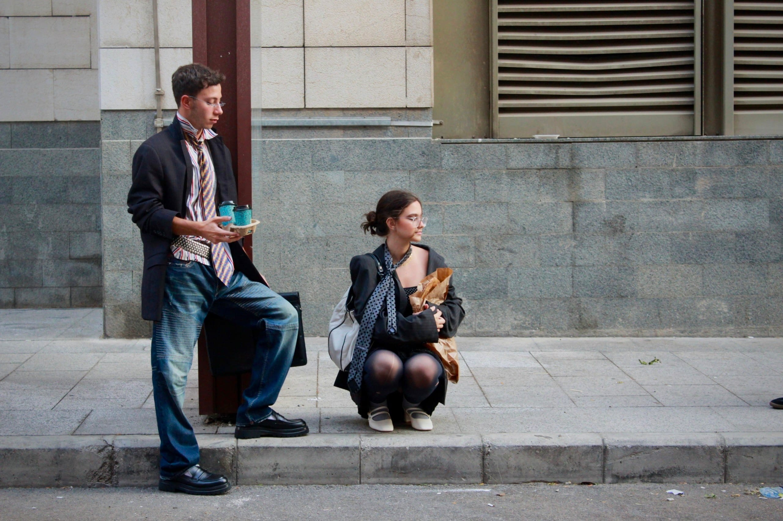 Model in a blazer and jeans standing on the kerb with coffees as a second model crouches alongside, clutching a paper bag