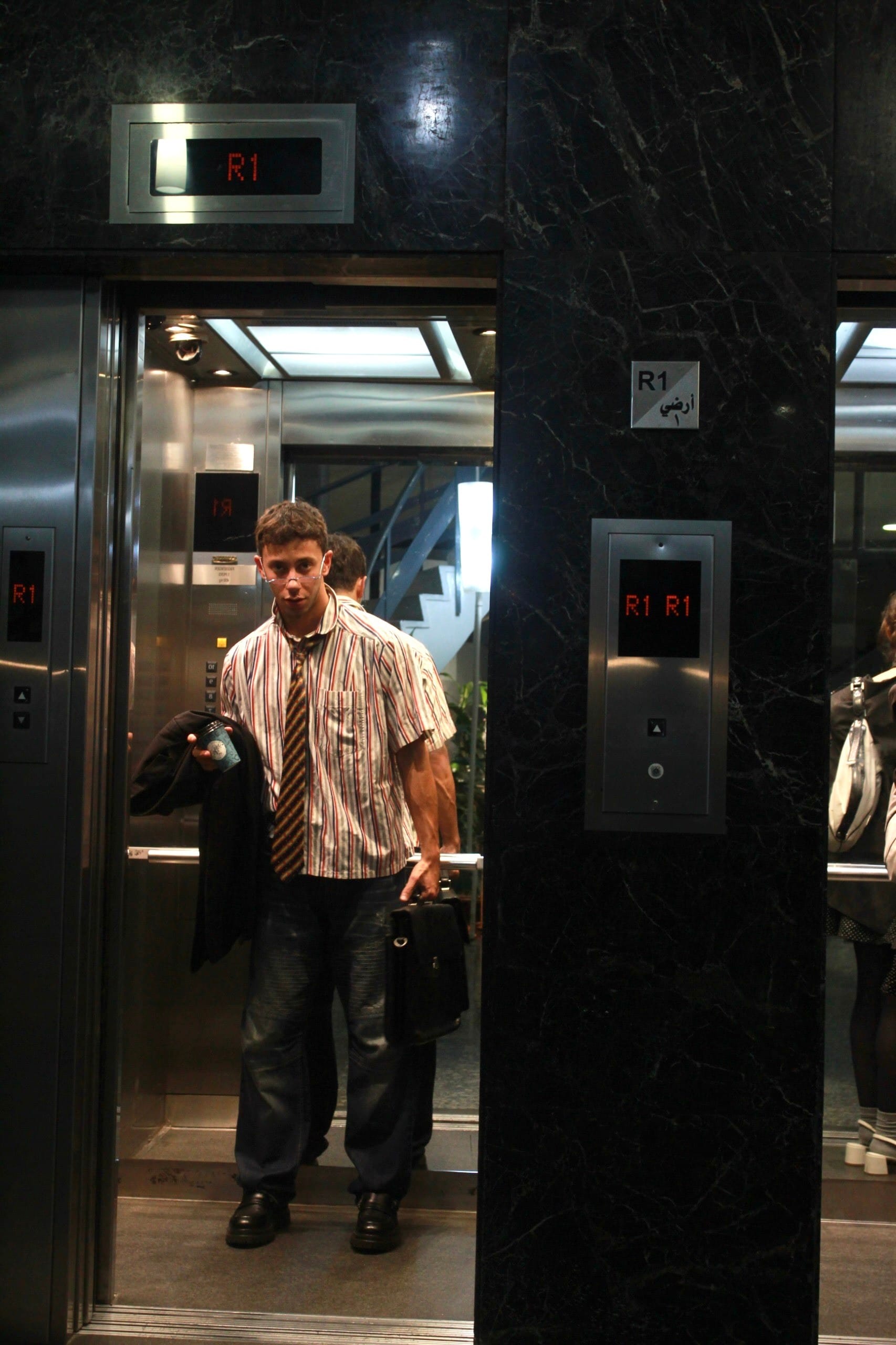 Model in a striped shirt and tie stepping out of a lift between black marble walls, jacket and briefcase in hand, another figure behind