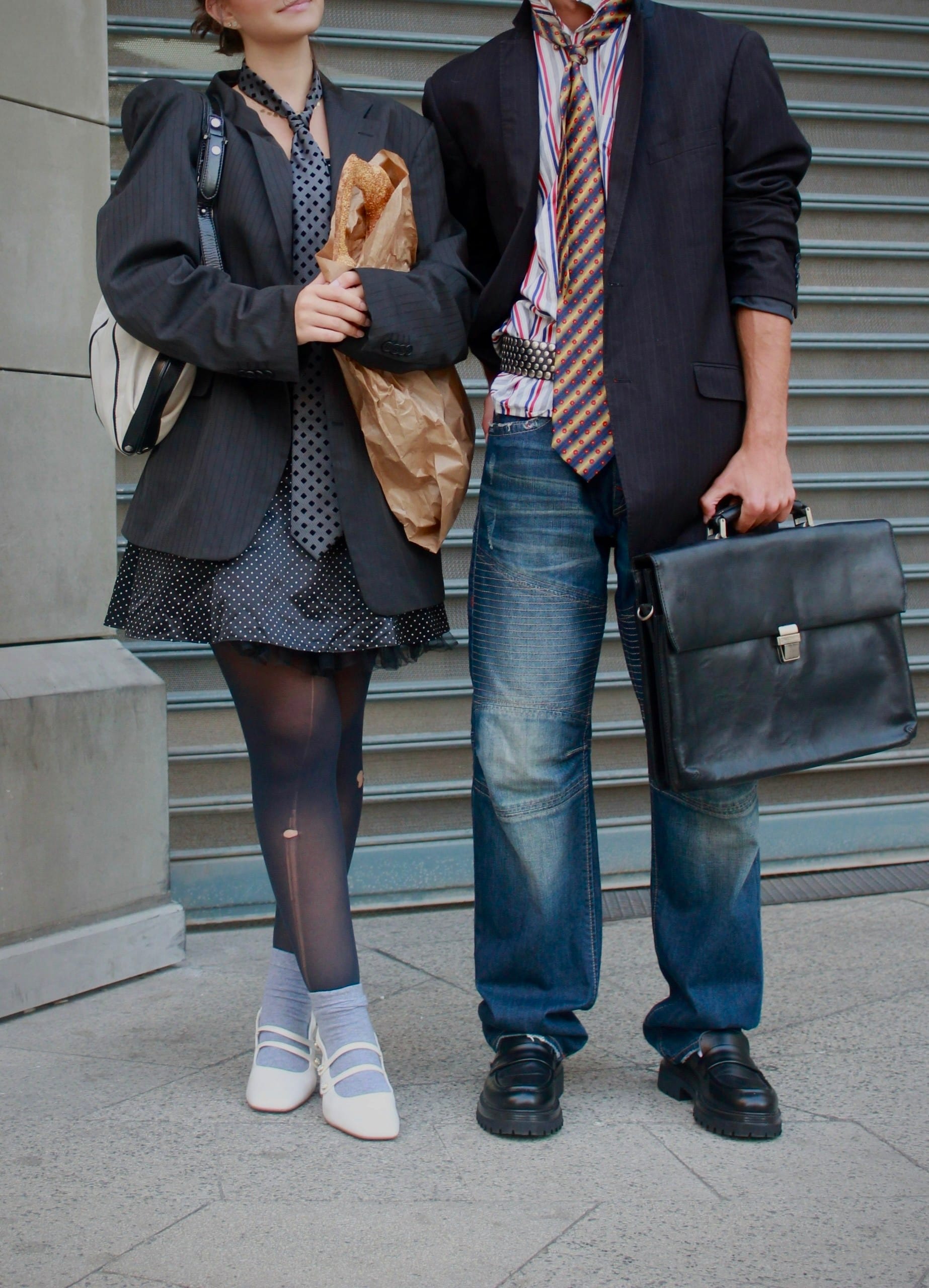 Two models cropped at the chin against a metal shutter, one hugging a paper bag of bread, the other in jeans holding a black briefcase