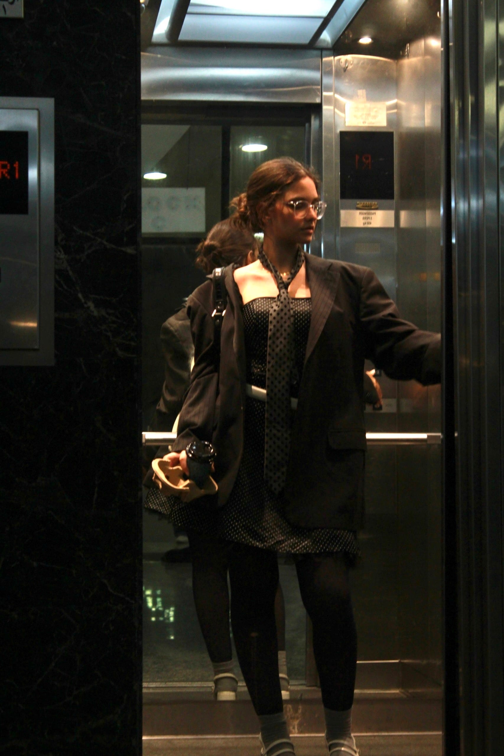 Model in glasses, a black blazer, polka-dot dress and skinny tie standing in a mirrored lift holding a coffee, reflected in the mirror behind