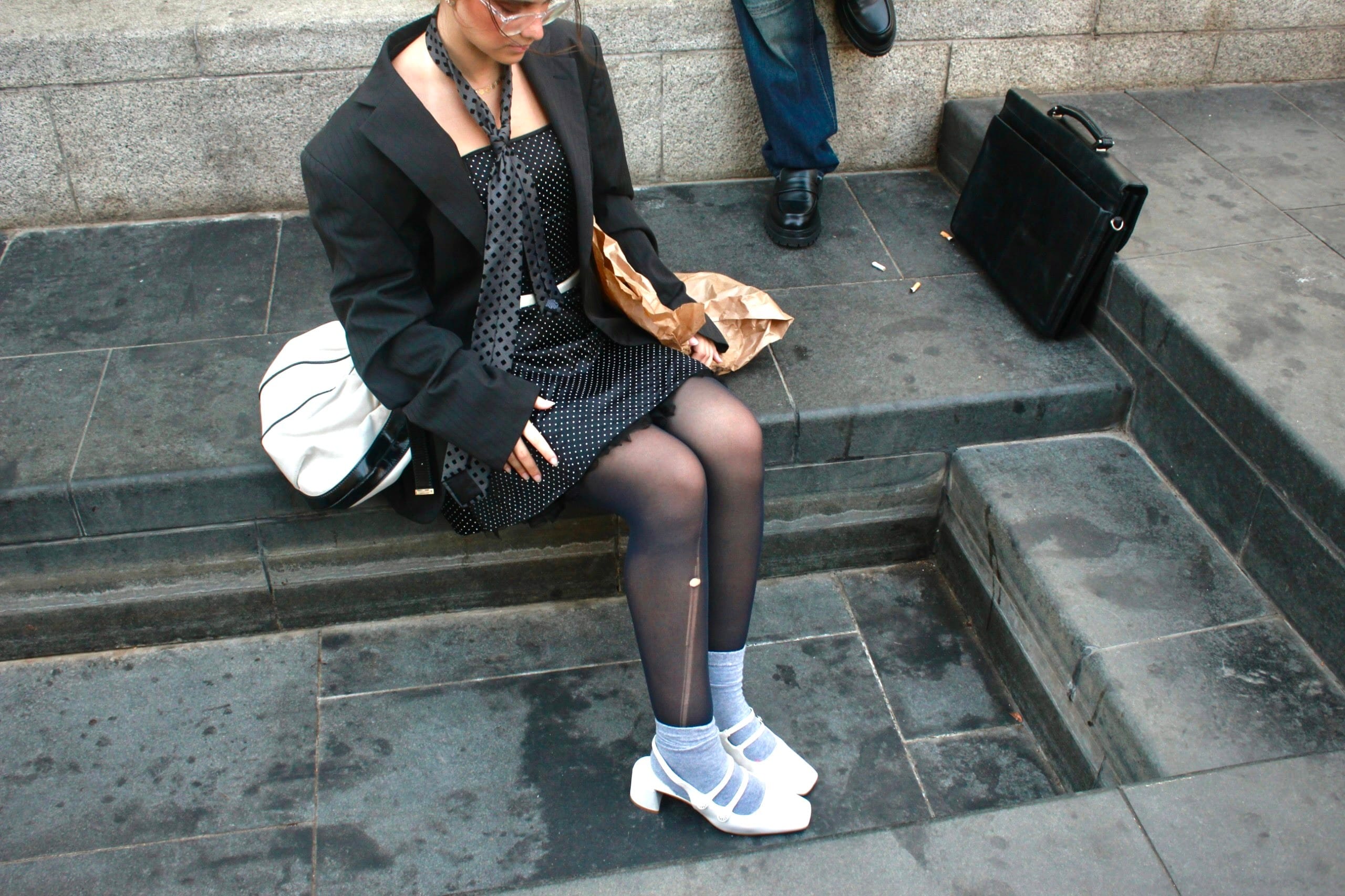 High-angle shot of a model seated on stone steps with head bowed, laddered black tights, grey socks and white Mary Jane heels, briefcase nearby