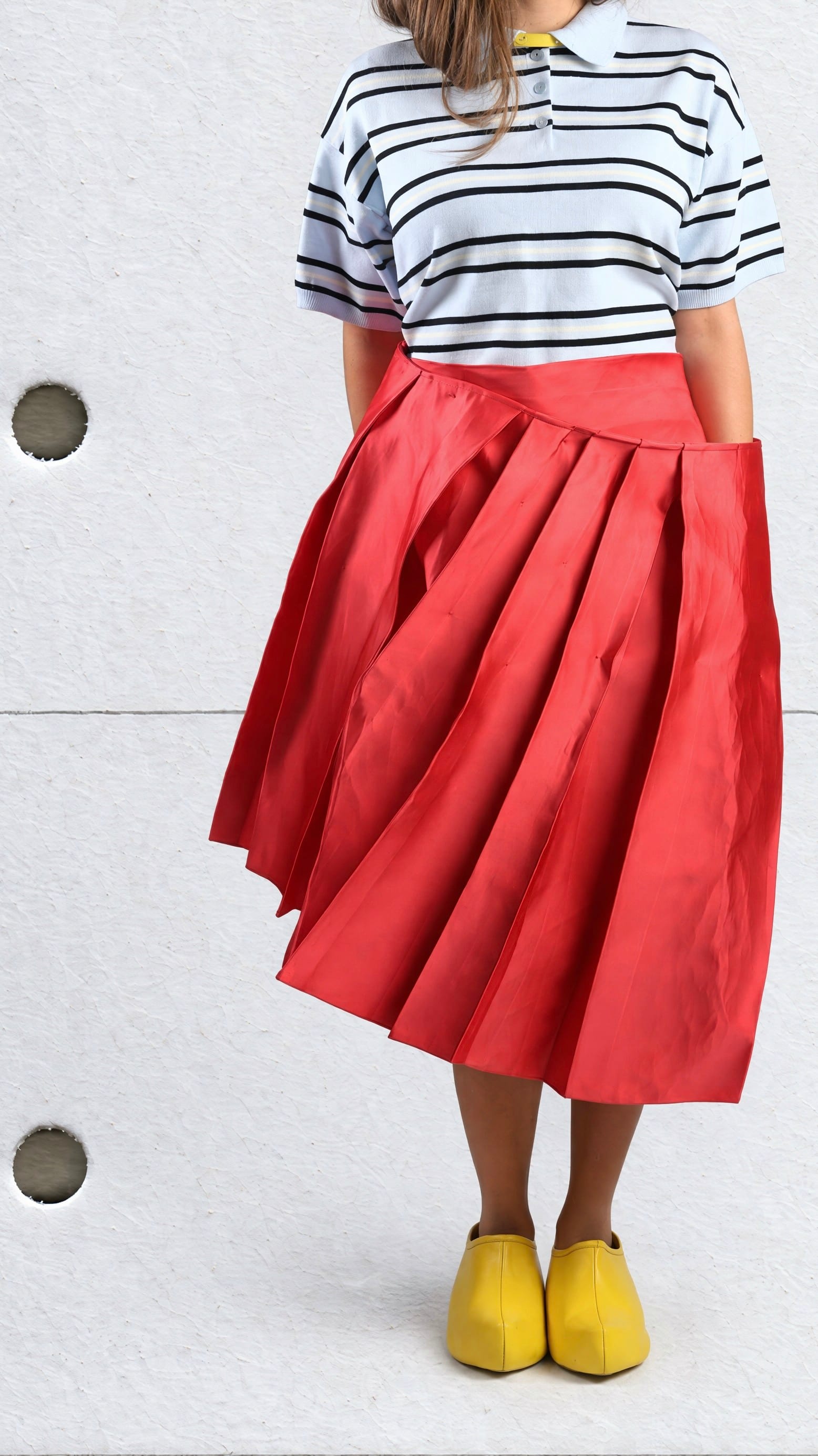 Chin-down view of a standing model in a black-striped pale blue polo and voluminous red pleated midi skirt, hand in pocket, yellow slip-on shoes