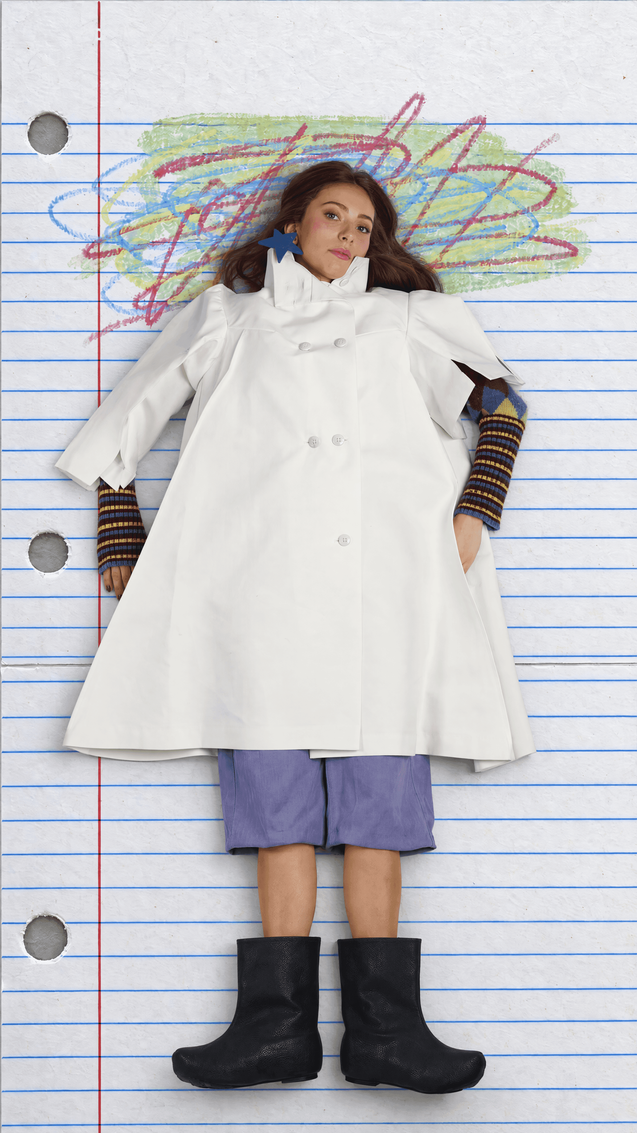 Overhead full-length view of a model lying on lined notebook paper under a crayon scribble in a white double-breasted coat, lavender shorts and black boots