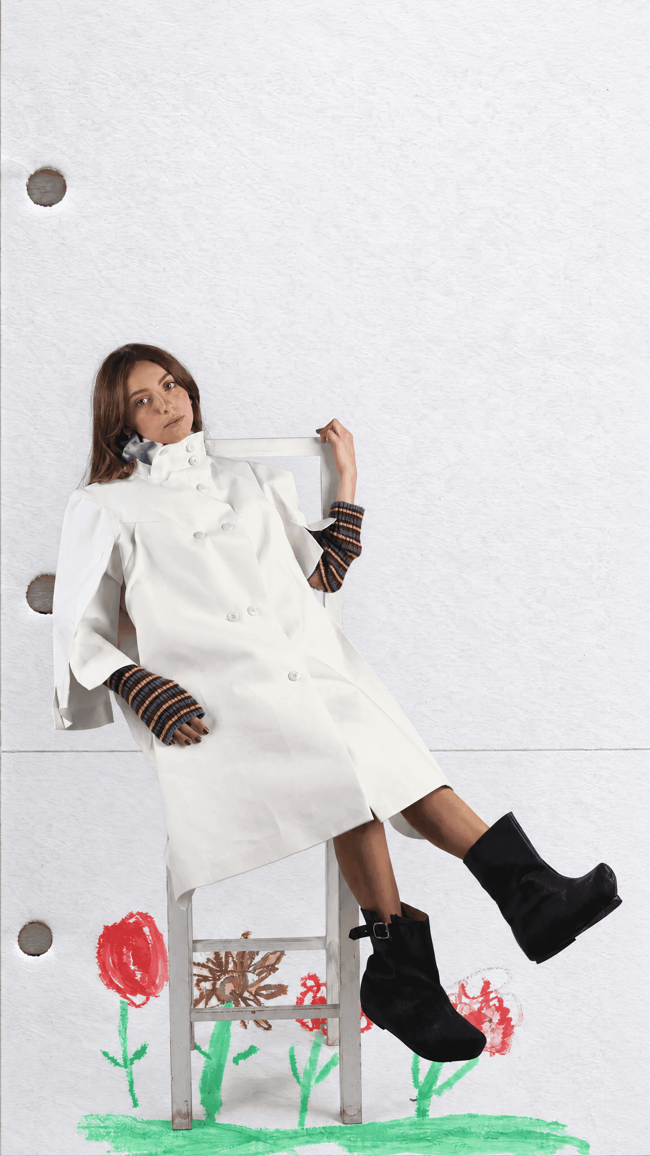 Model lounging on a white chair in a white double-breasted coat, striped fingerless gloves and black boots, one leg raised above crayon-drawn flowers