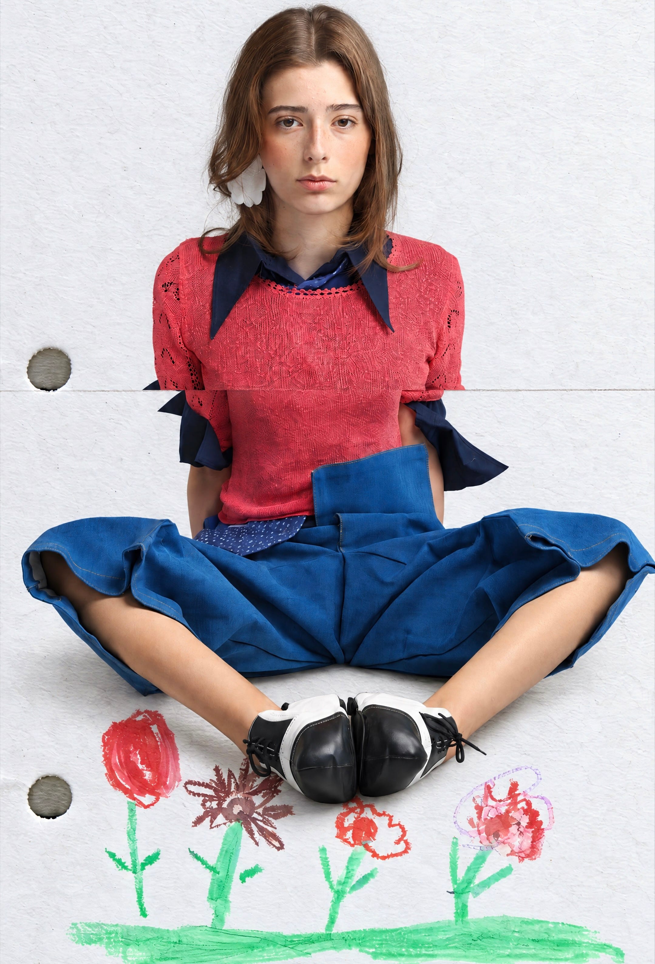 Model seated with feet together in a coral openwork knit top over a navy pointed-collar shirt and voluminous denim trousers, crayon flowers below