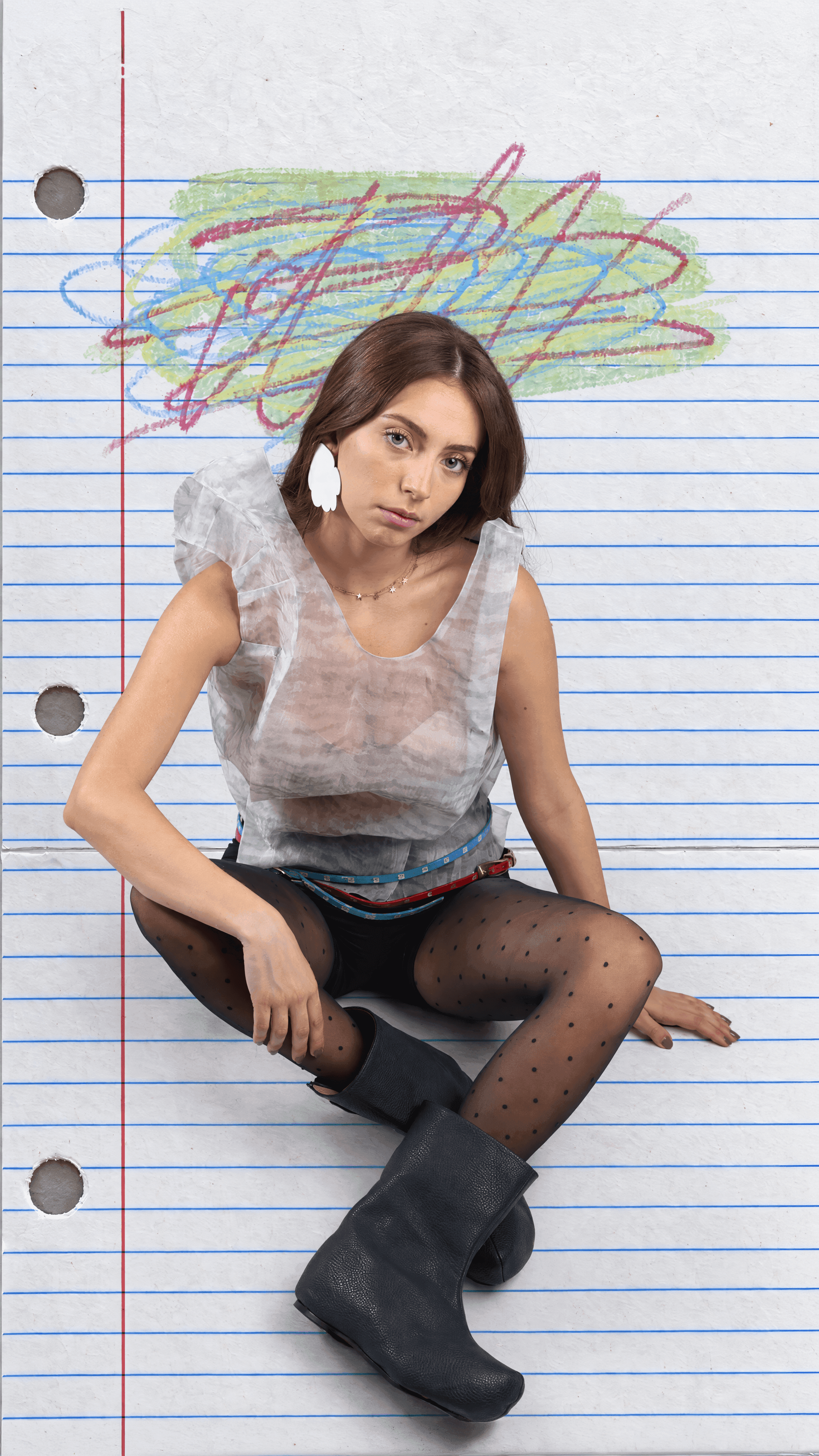 Model seated on lined notebook paper under a crayon scribble in a sheer sleeveless top, black shorts, polka-dot tights and black boots