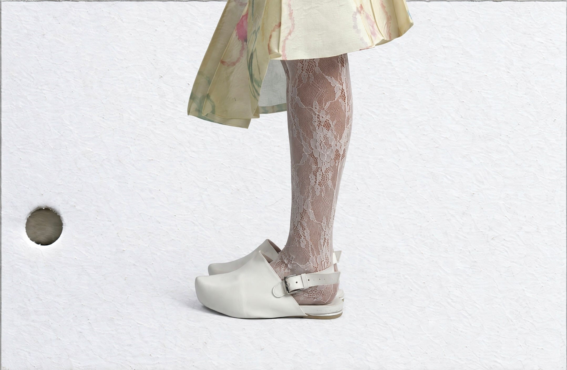 Side-view detail of a lower leg in white floral lace tights and a white buckled clog beneath a cream flower-print dress hem