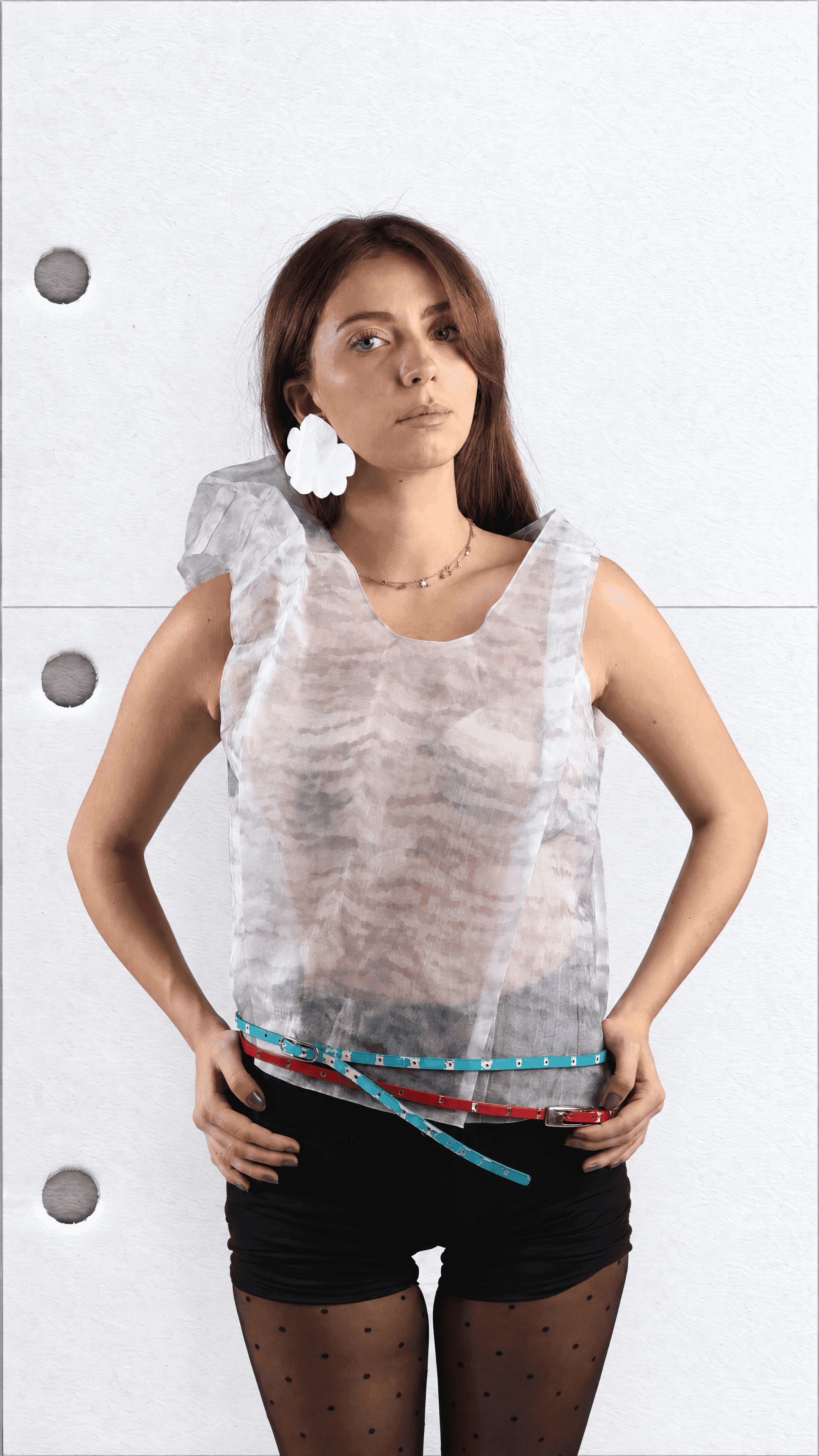 Thigh-up view of a model with hands on hips in a sheer sleeveless top with a crumpled shoulder, teal and red belts and black shorts
