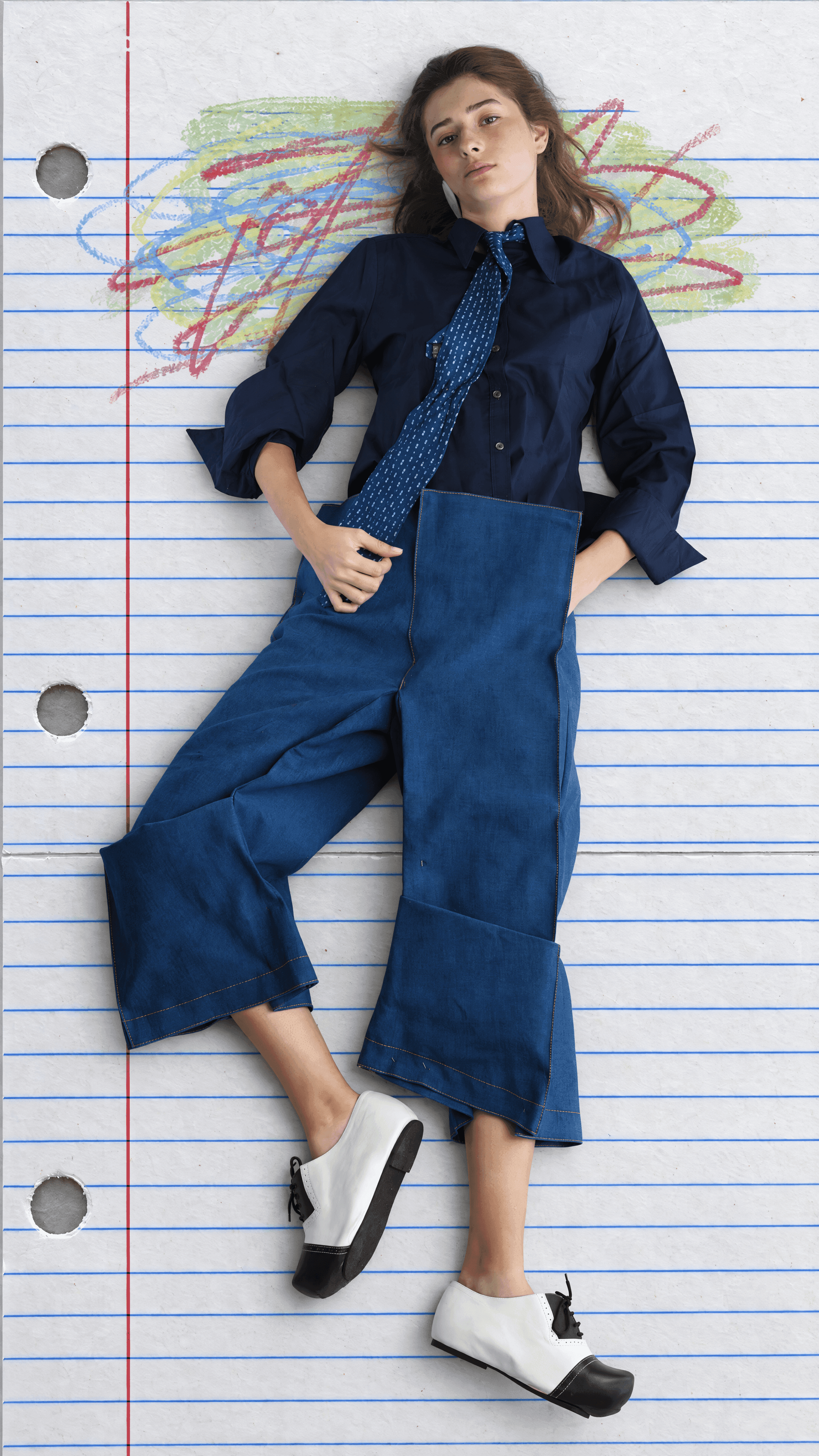 Overhead view of a model lying on lined notebook paper in a navy shirt, blue print tie and wide cropped denim trousers with deep cuffs