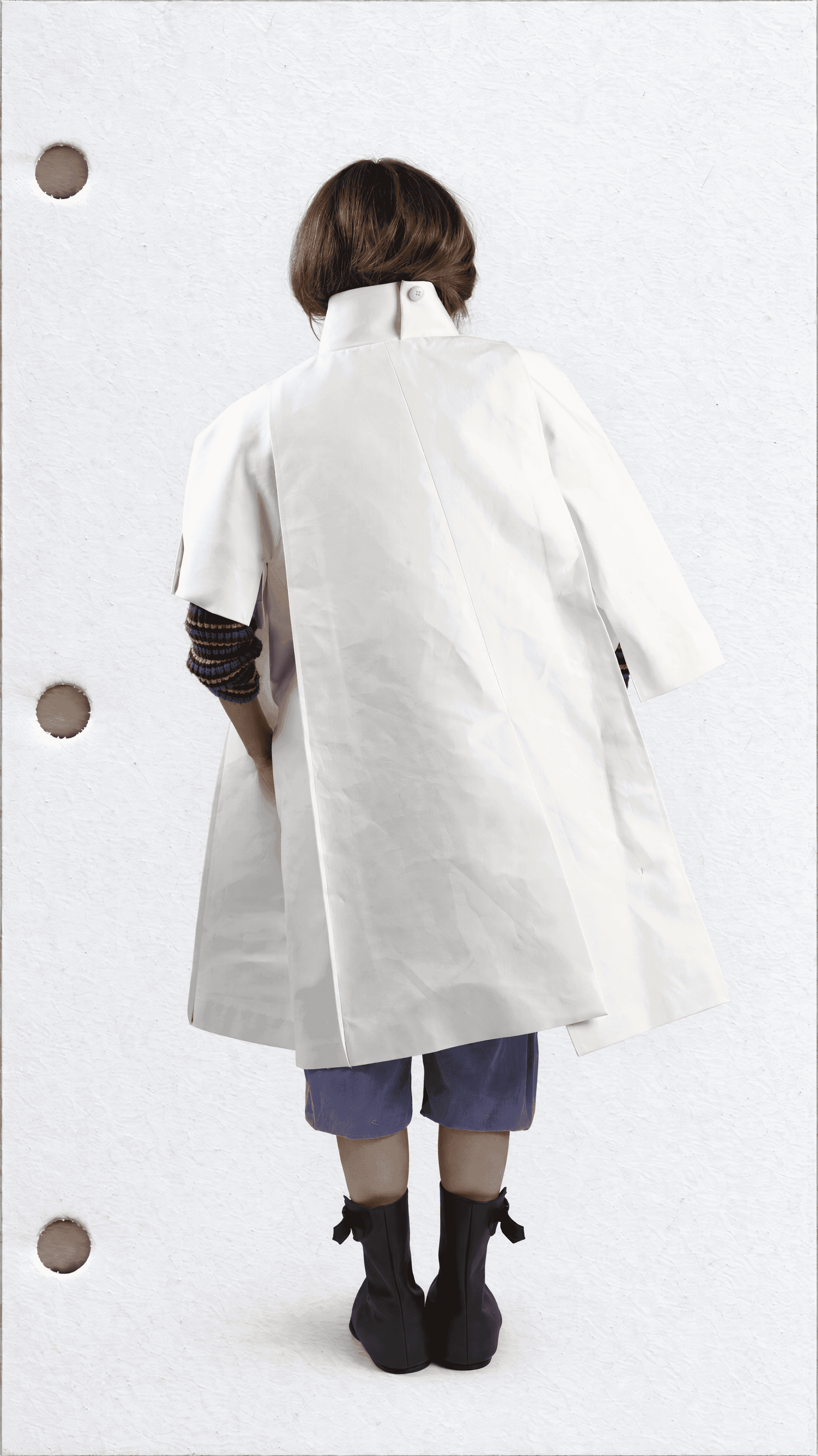 Full-length back view of a model in a white A-line coat, showing its tall collar and layered back panel, with lavender shorts and black boots