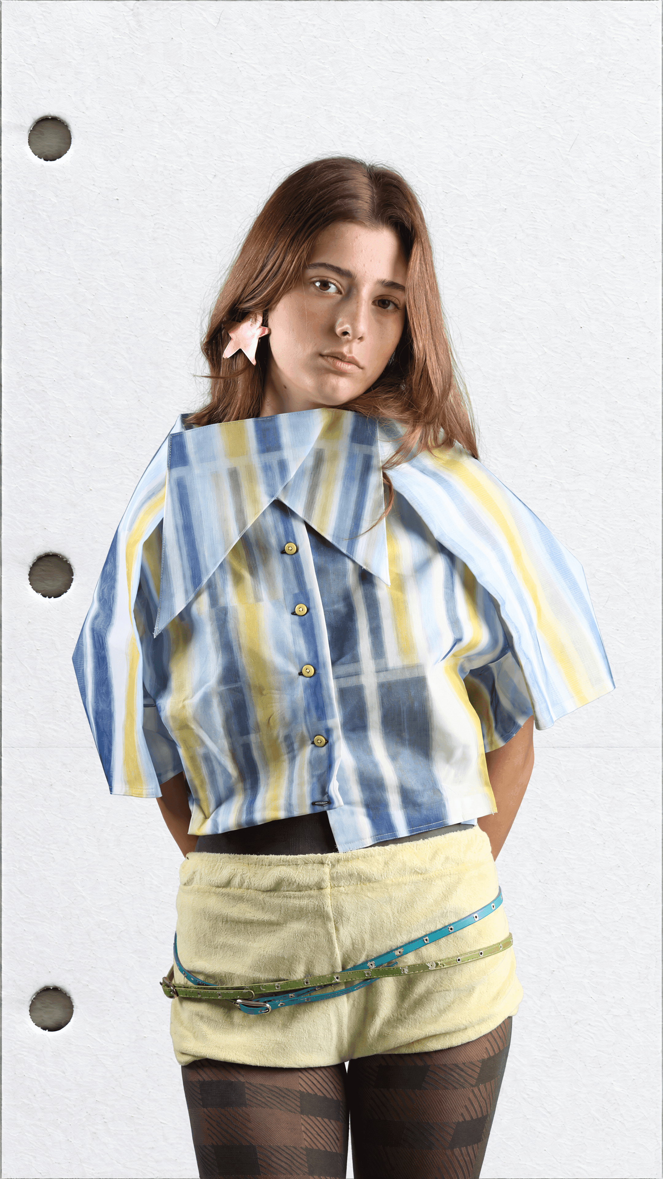 Thigh-up view of a standing model in a blue and yellow striped shirt with a wide pointed collar, pale yellow shorts and two skinny belts
