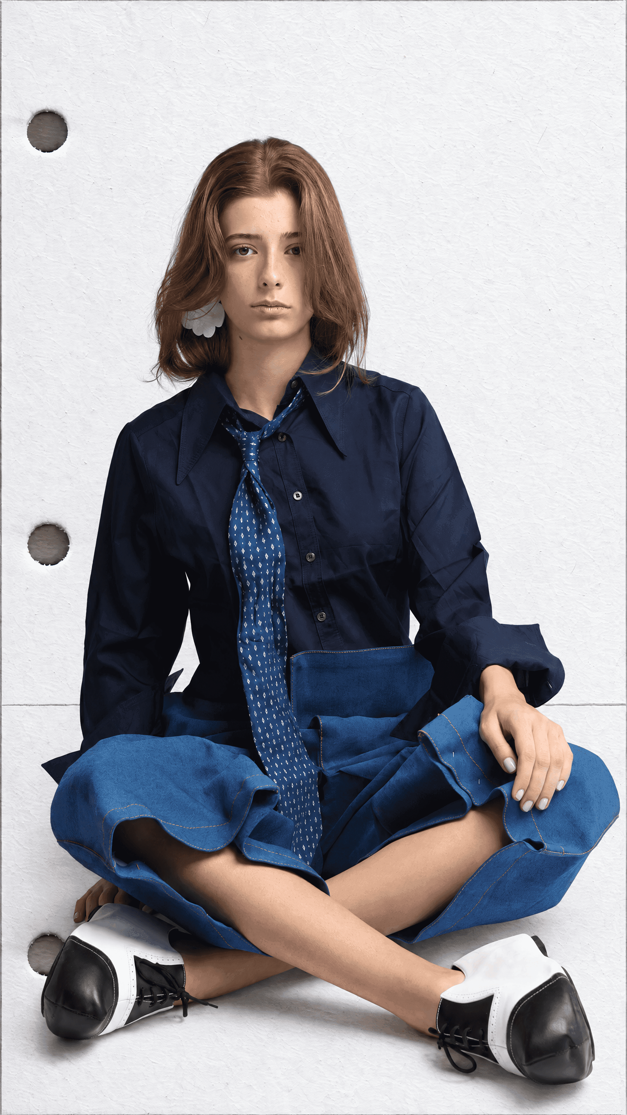 Model sitting cross-legged on hole-punched white paper in a navy pointed-collar shirt, blue print tie, voluminous denim trousers and black-and-white lace-ups