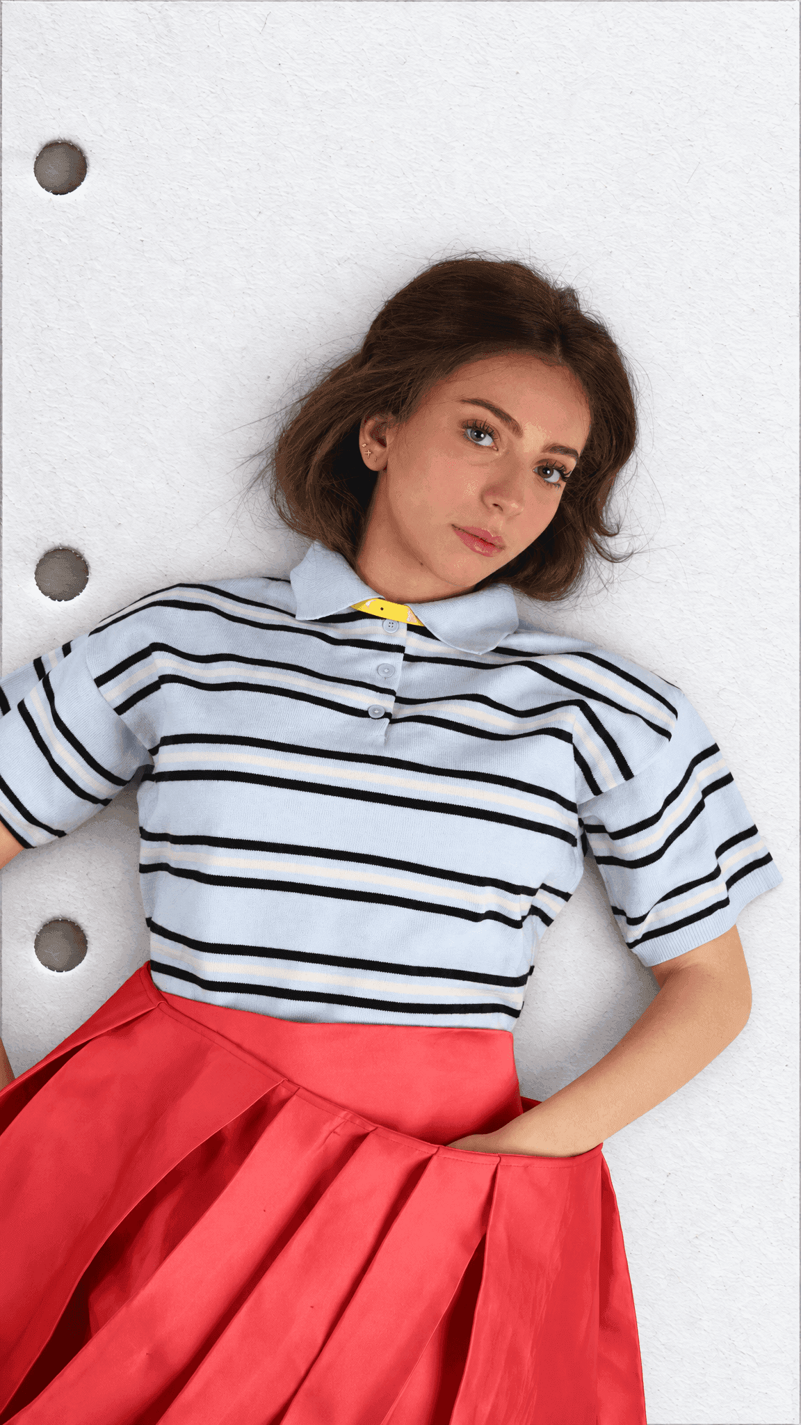 Waist-up overhead view of a model lying down in a black-striped pale blue polo with a yellow collar detail and red pleated skirt