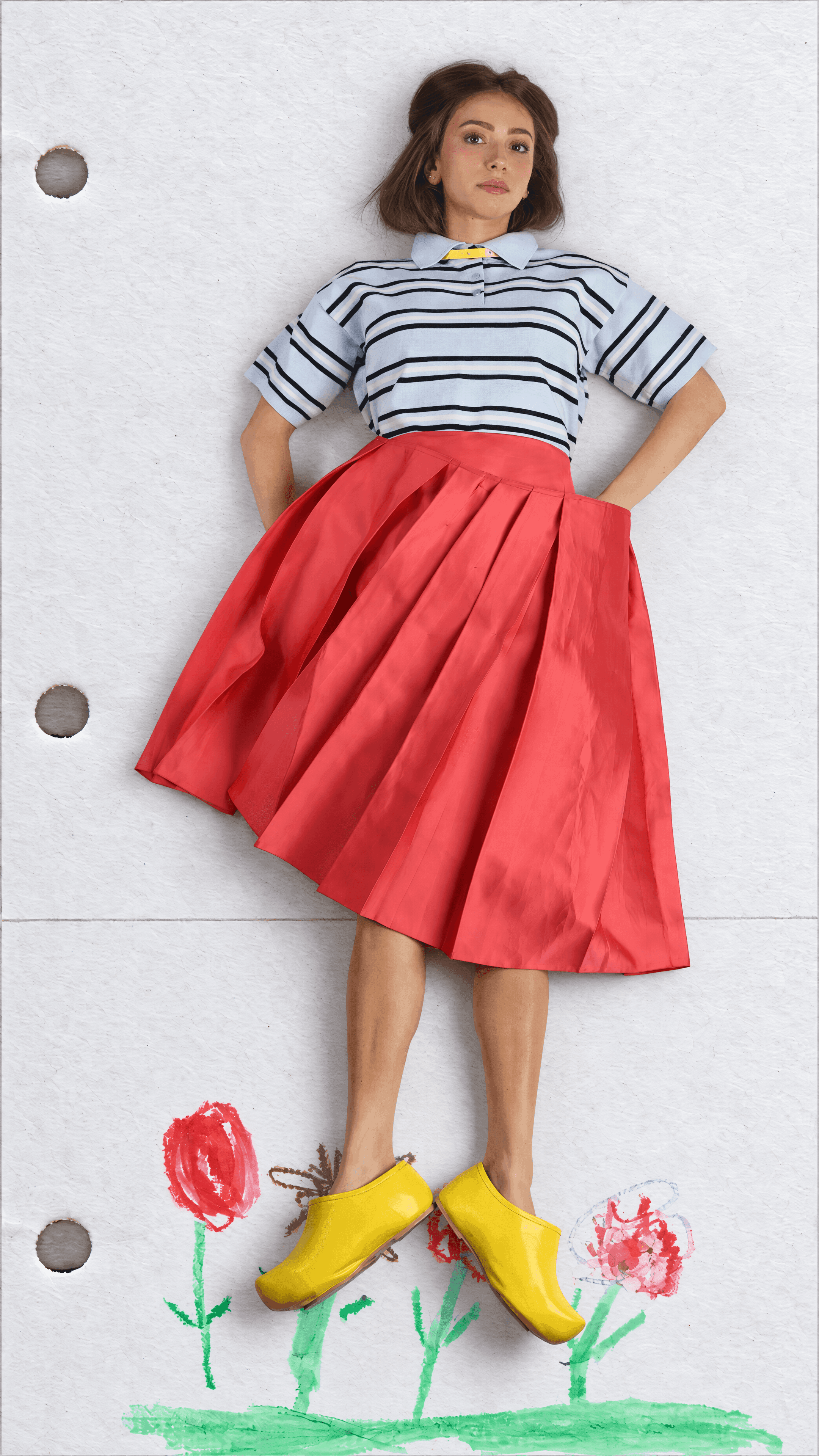 Full-length view of a model, hands on hips, in a striped pale blue polo and full red pleated skirt with yellow shoes above crayon flowers