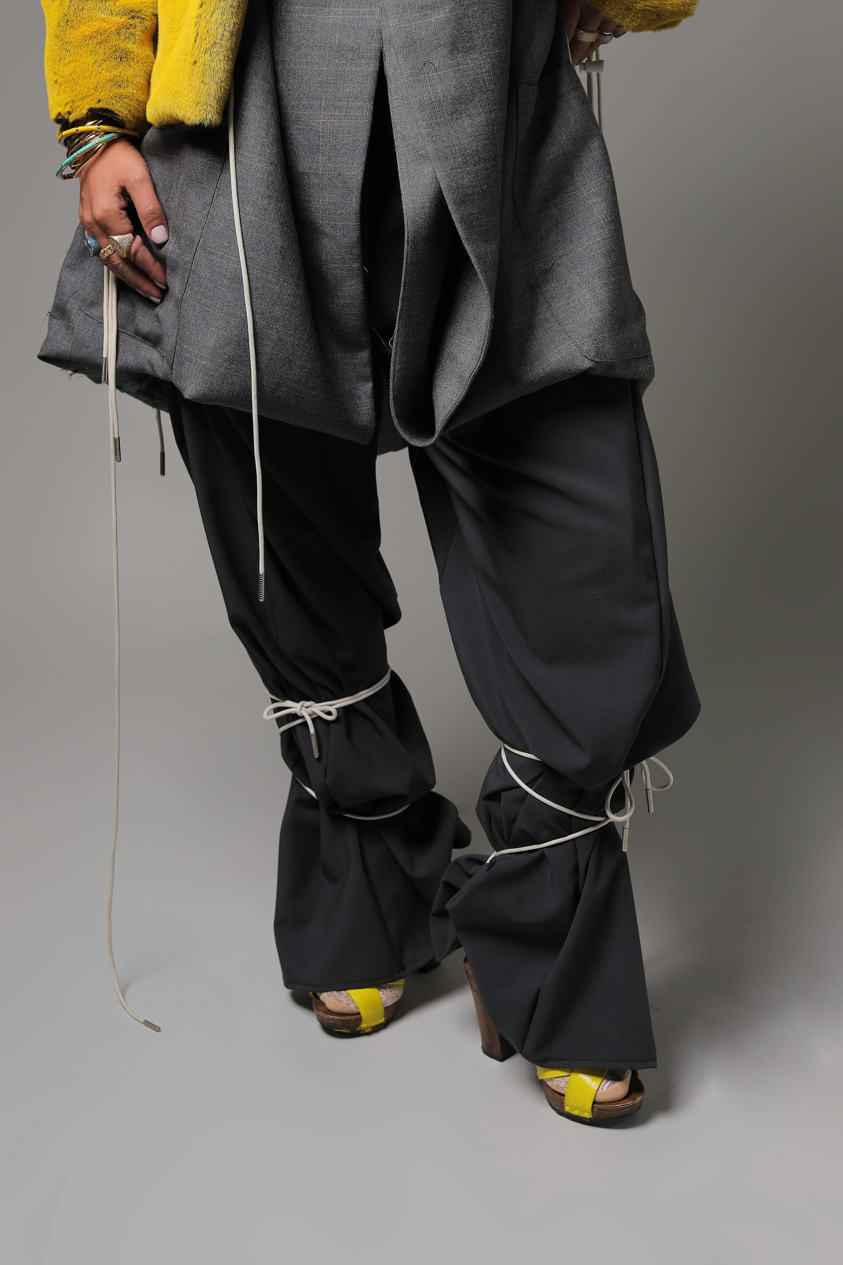 Lower-body detail of charcoal trousers cinched with white cords at the knees, a drawstring grey blazer above and yellow-strapped heels below
