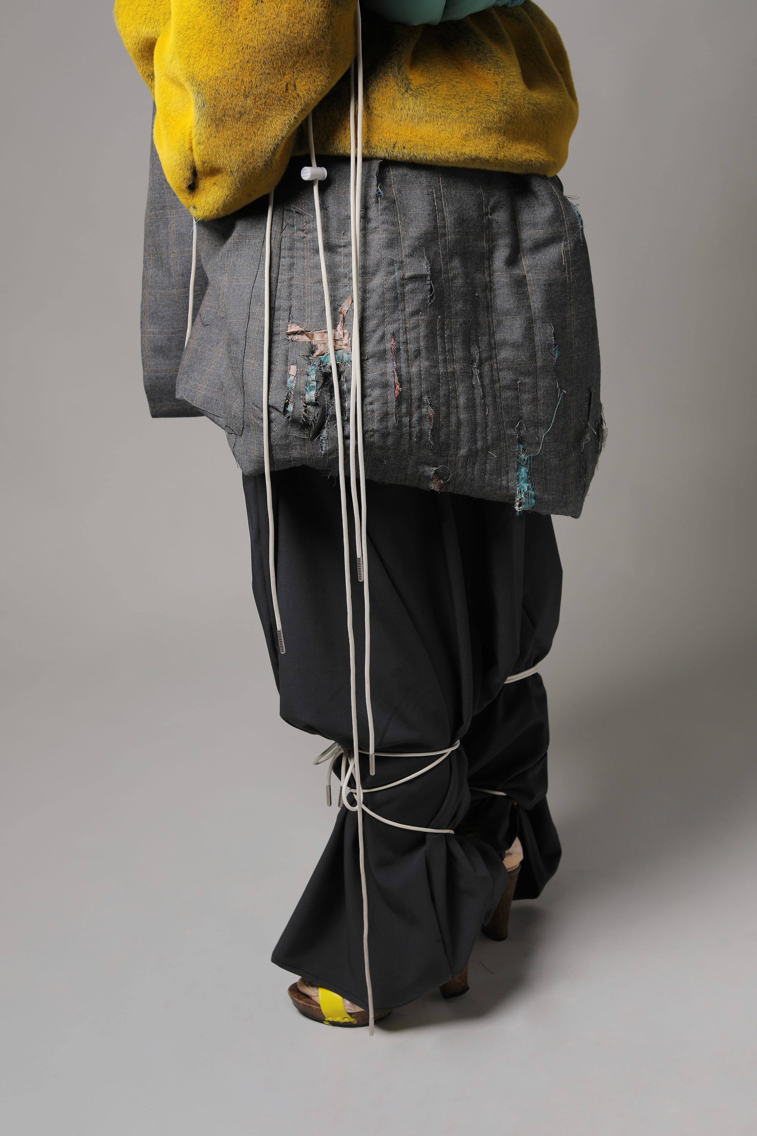 Side view of a distressed grey blazer hem with frayed patches and long drawstrings above charcoal trousers tied at the knees