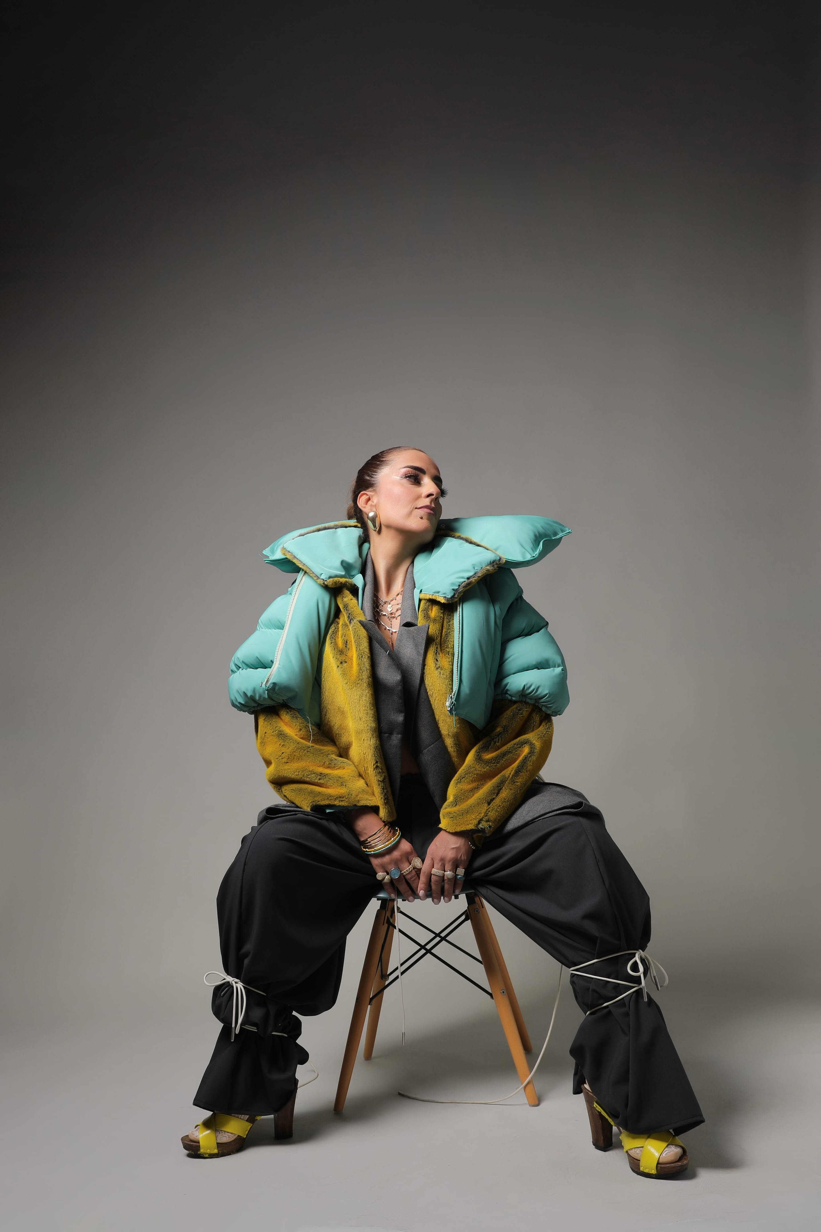 Full-length shot of the model seated on a wooden stool, legs apart, in the mint puffer, yellow plush jacket and cord-tied wide trousers
