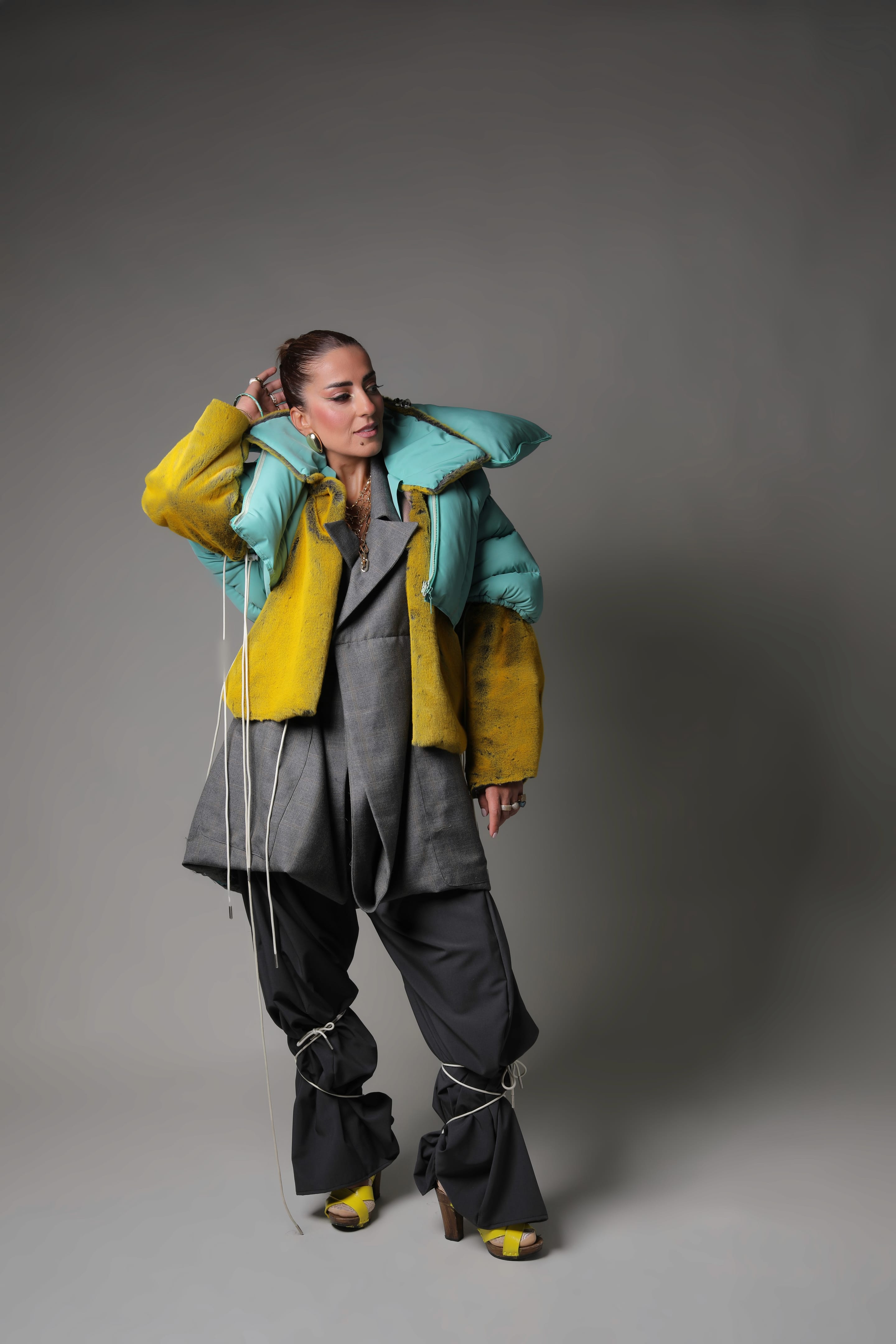Full-length shot of the model, hand to head, in a mint padded collar and yellow plush jacket over a grey blazer and cord-tied trousers