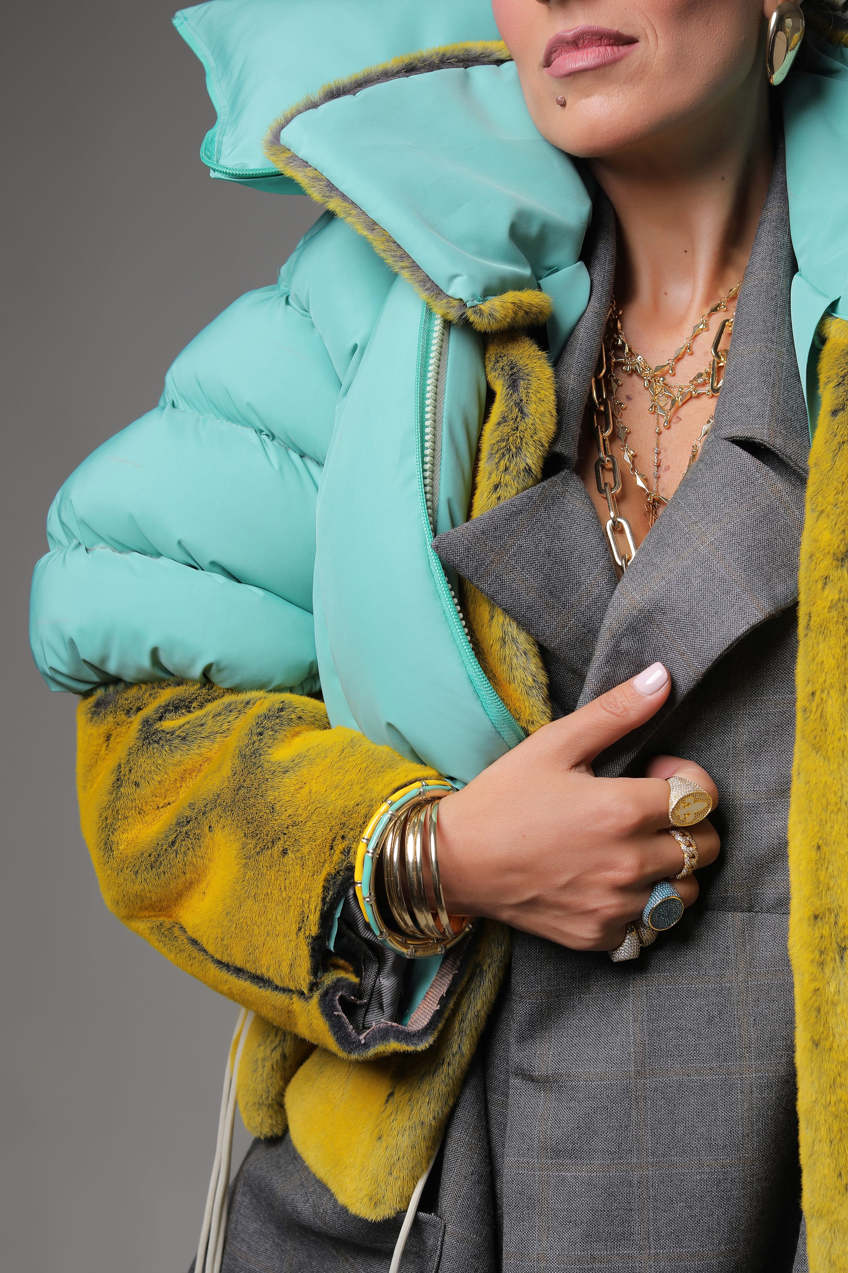 Close-up of the mint puffer's padded collar and yellow plush sleeve over a grey blazer lapel, a ringed hand at the chest